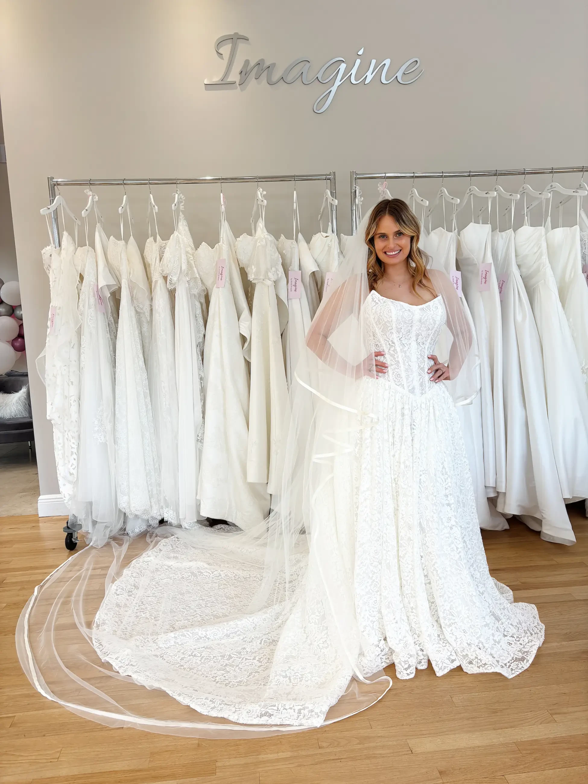 A woman smiles in a bridal shop, wearing a lace wedding gown with a long train. Behind her, a row of white dresses hangs under the word 