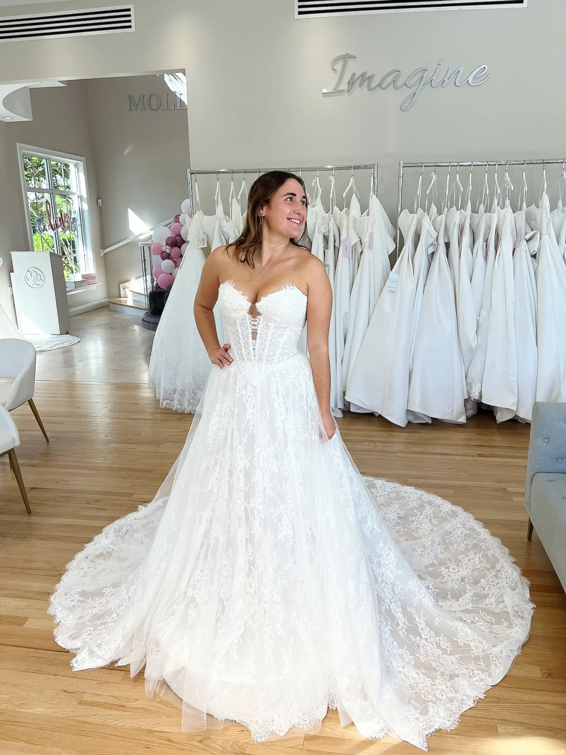 A woman joyfully models a lacy white wedding dress with a flowing train in a bridal shop. Racks of dresses and 