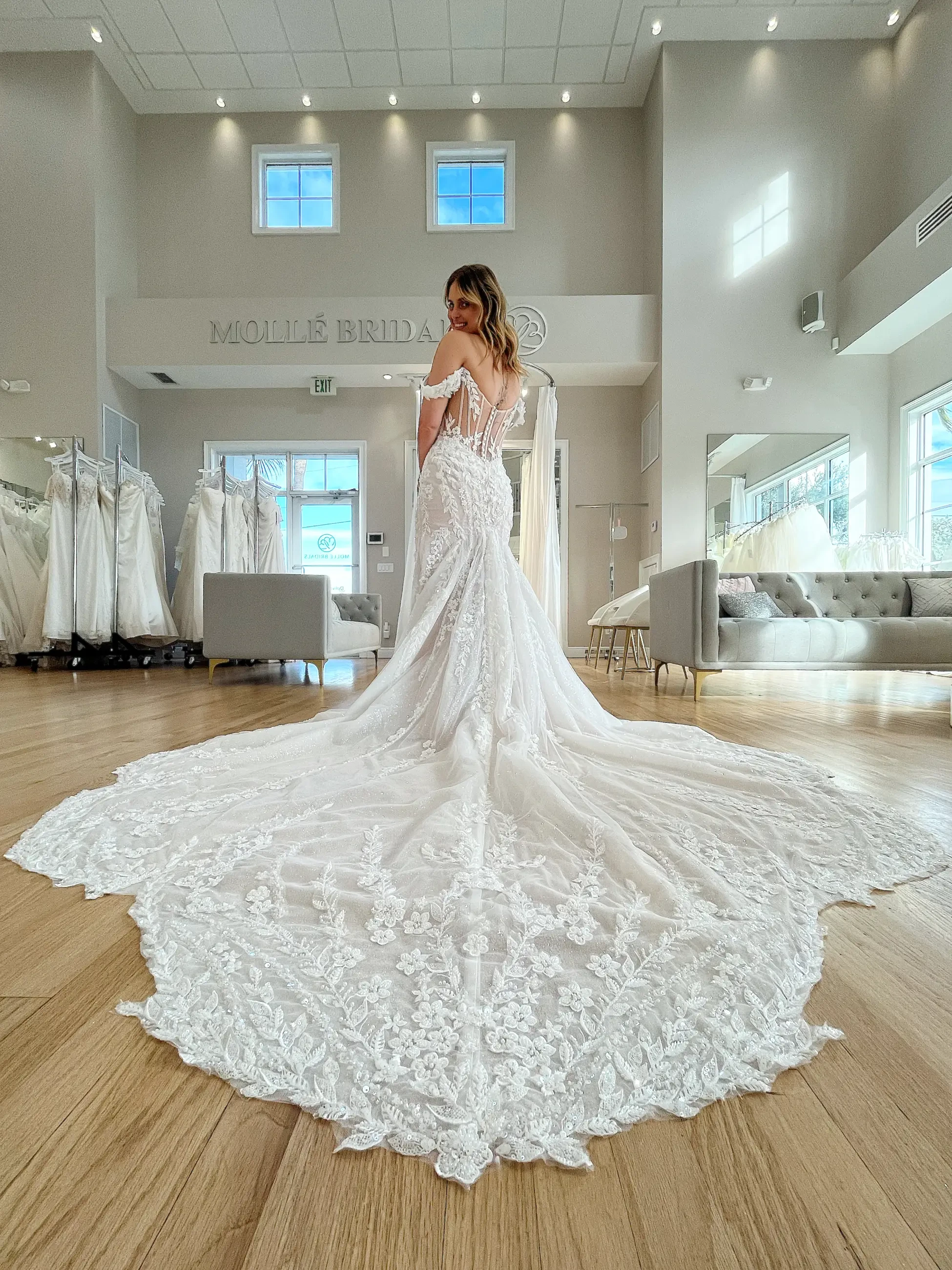A bride in a flowing lace gown stands in a bridal boutique, her long train elegantly spread across the wooden floor. Soft lighting creates a dreamy ambiance.