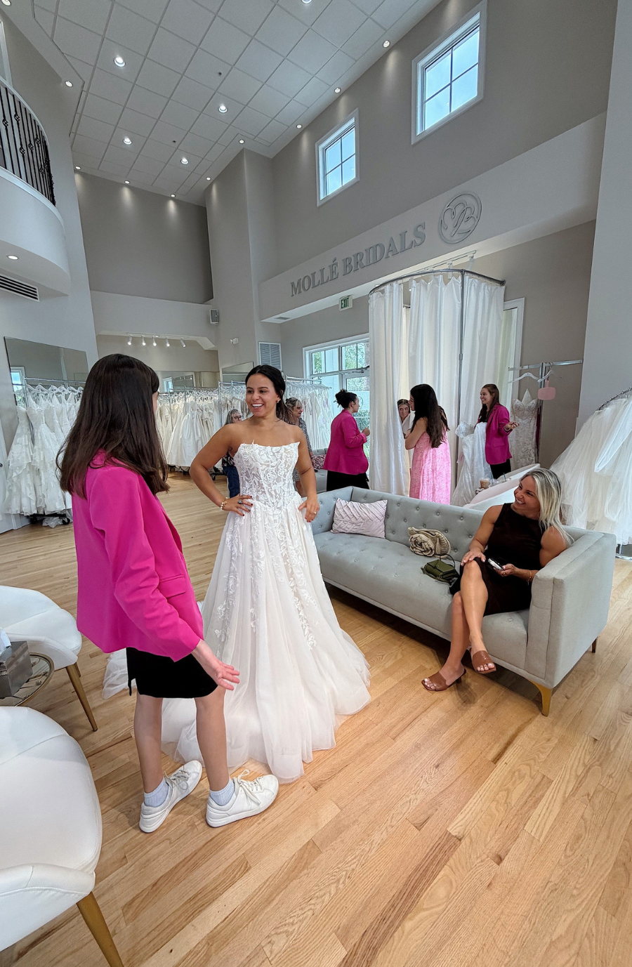  A bride trying on a wedding dress in a bridal shop, with a girl in casual attire standing nearby and several women in the background.