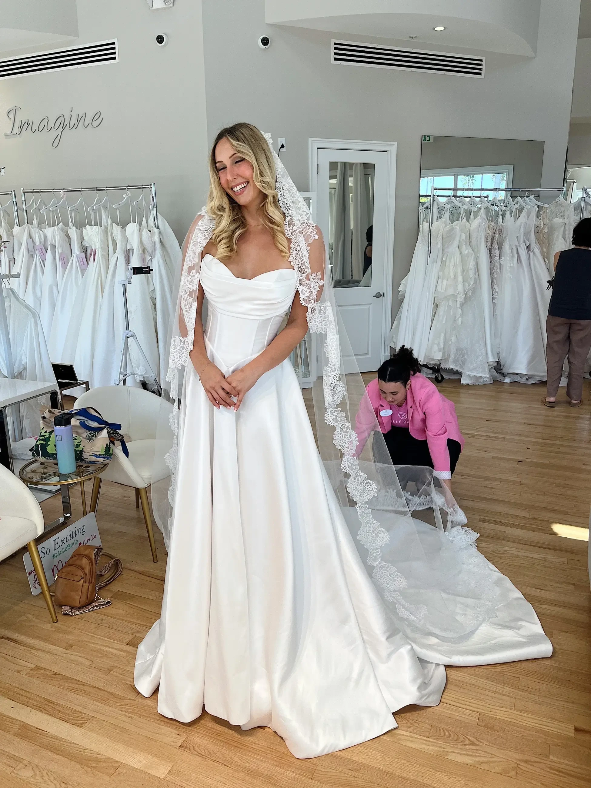 A bride stands in a bridal shop wearing a white wedding gown with a sweetheart neckline and long lace veil, while two women assist her in the background.