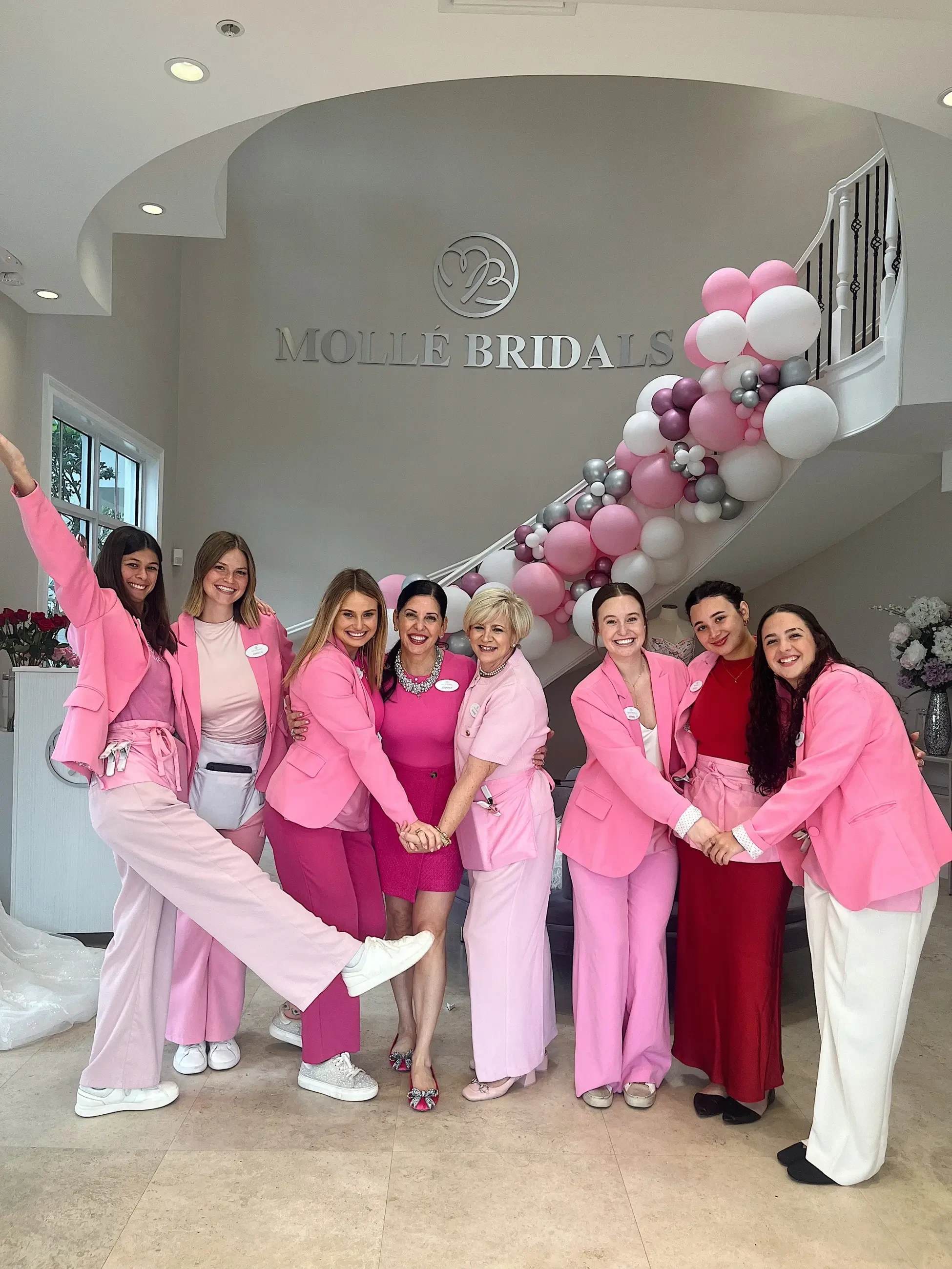 A group of women wearing pink outfits posing together in a bridal shop decorated with balloons.