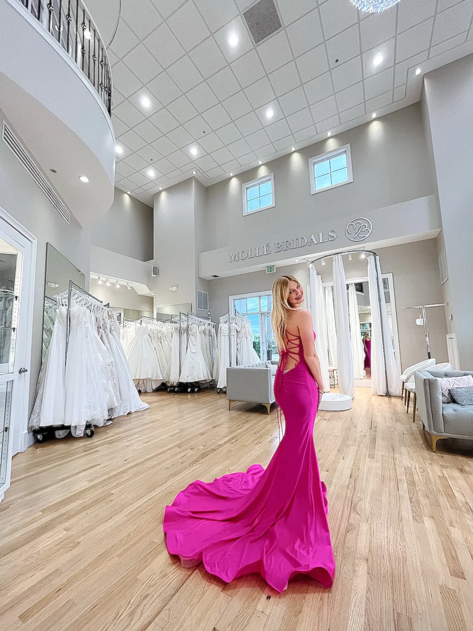 A woman in a vibrant pink gown stands in a bridal shop, with wedding dresses displayed around her. The bright and elegant interior features wooden flooring and large windows.