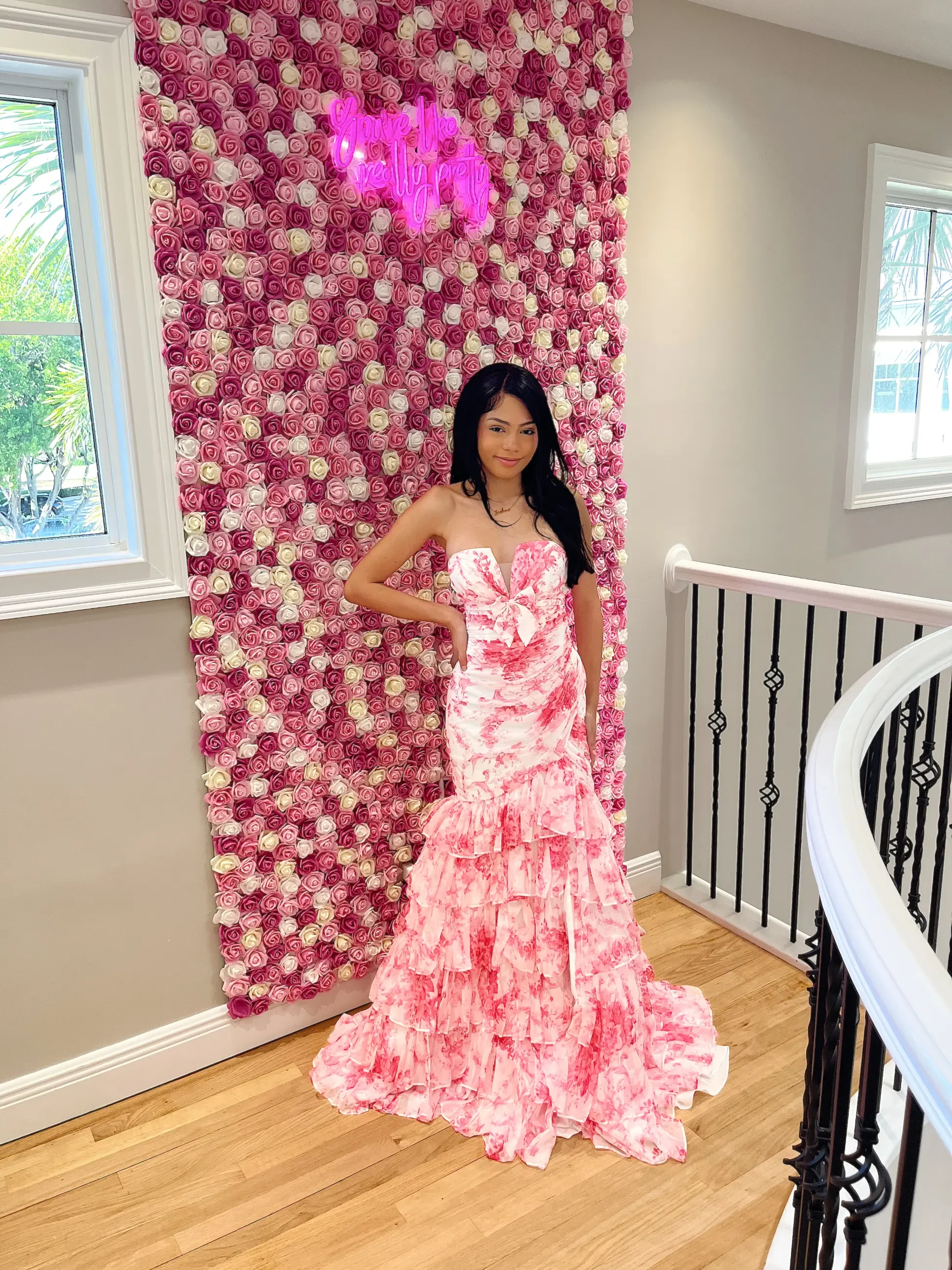 A woman in a pink and white floral dress poses confidently in front of a pink rose wall with a neon sign. The scene feels elegant and cheerful.