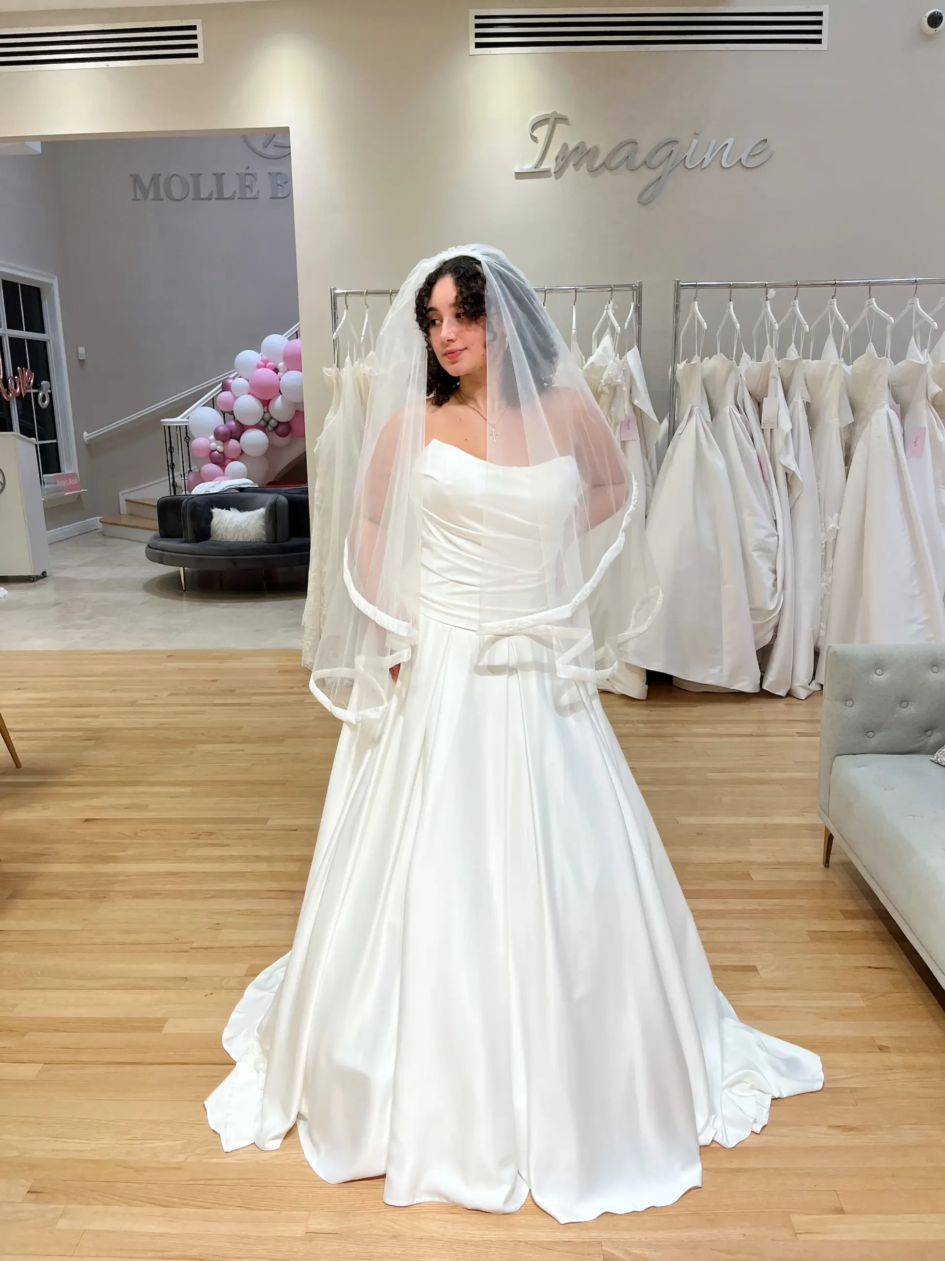 A bride in a white gown with a strapless bodice and long veil stands on wood flooring in a bridal shop. Behind her are racks of dresses and a pink balloon display.