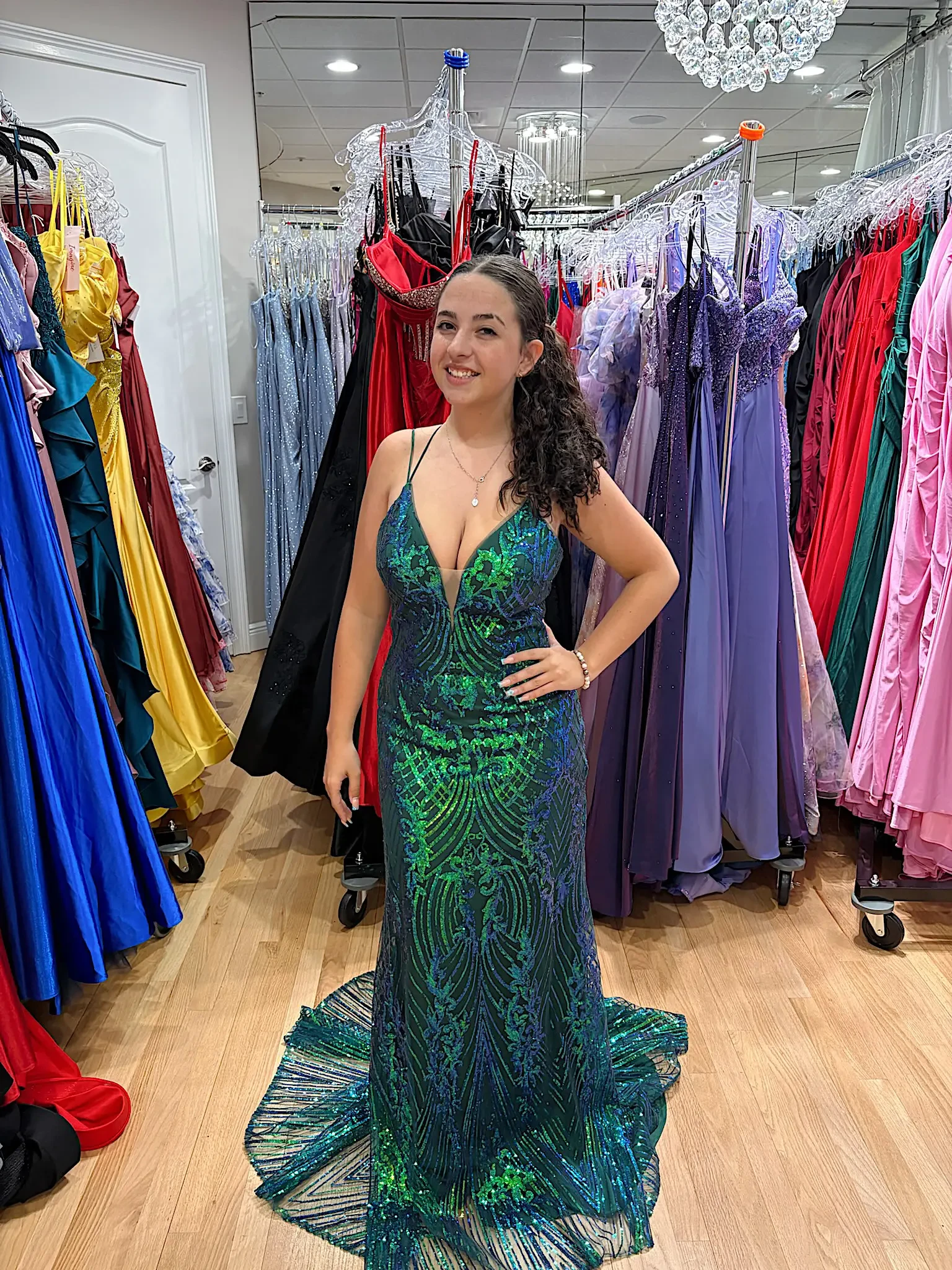A young woman wearing a sparkling green dress stands in a room filled with various colorful gowns on display.