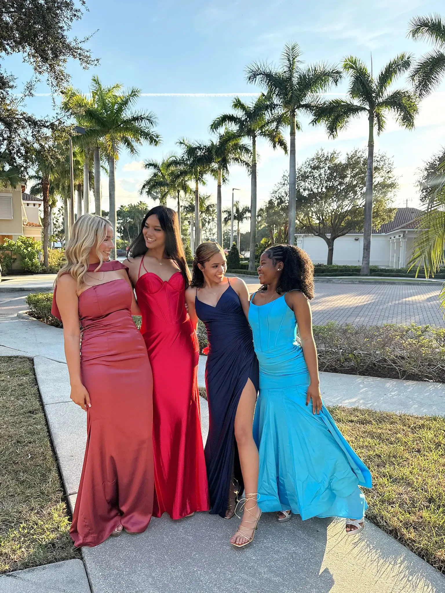 Four women in formal gowns, each a different vivid color, share a joyful moment outdoors. Palm trees and soft sunlight create a tropical ambiance.