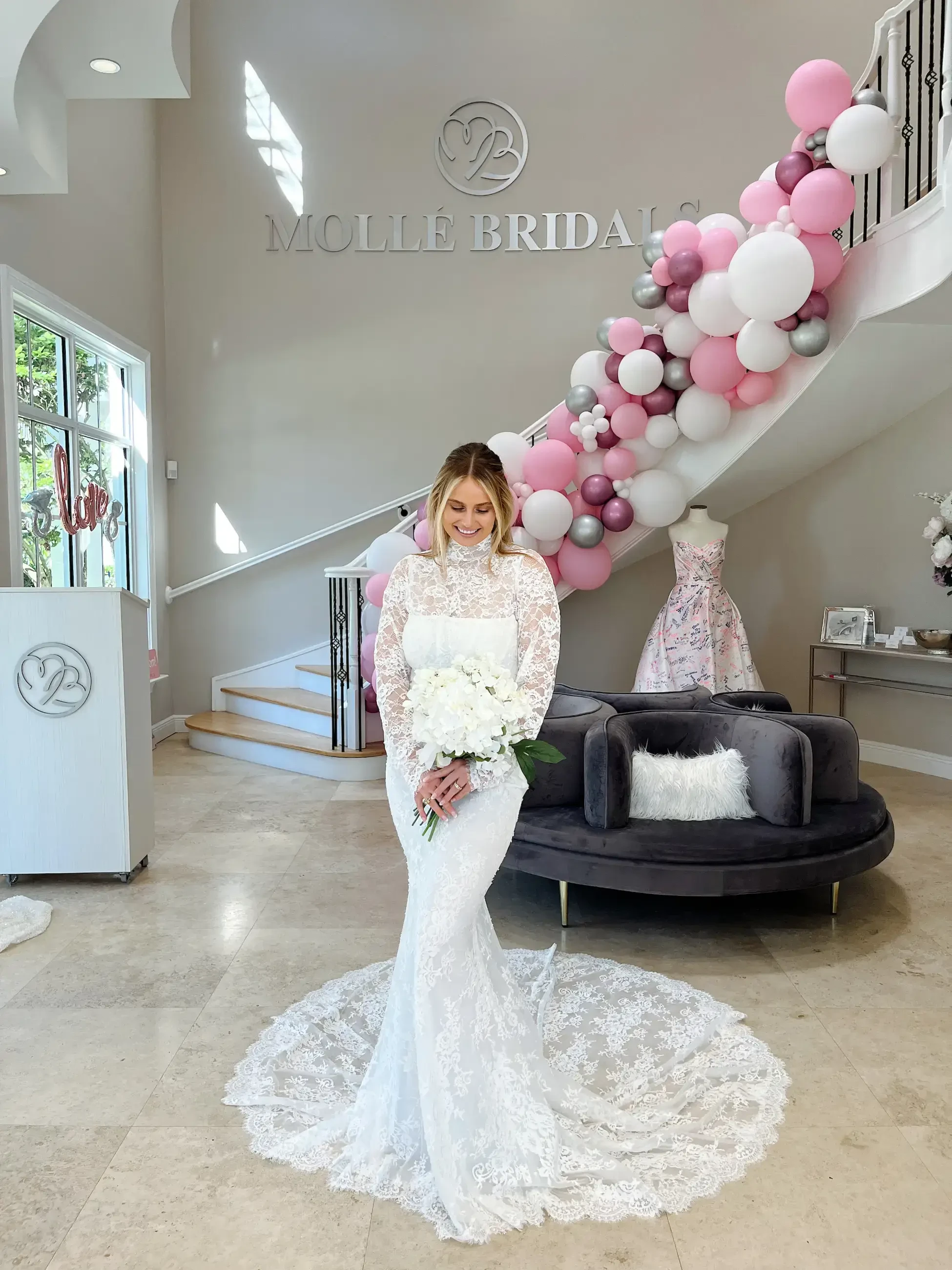 A woman in a strapless white wedding dress with a flared skirt stands on a pedestal in a bridal shop.
