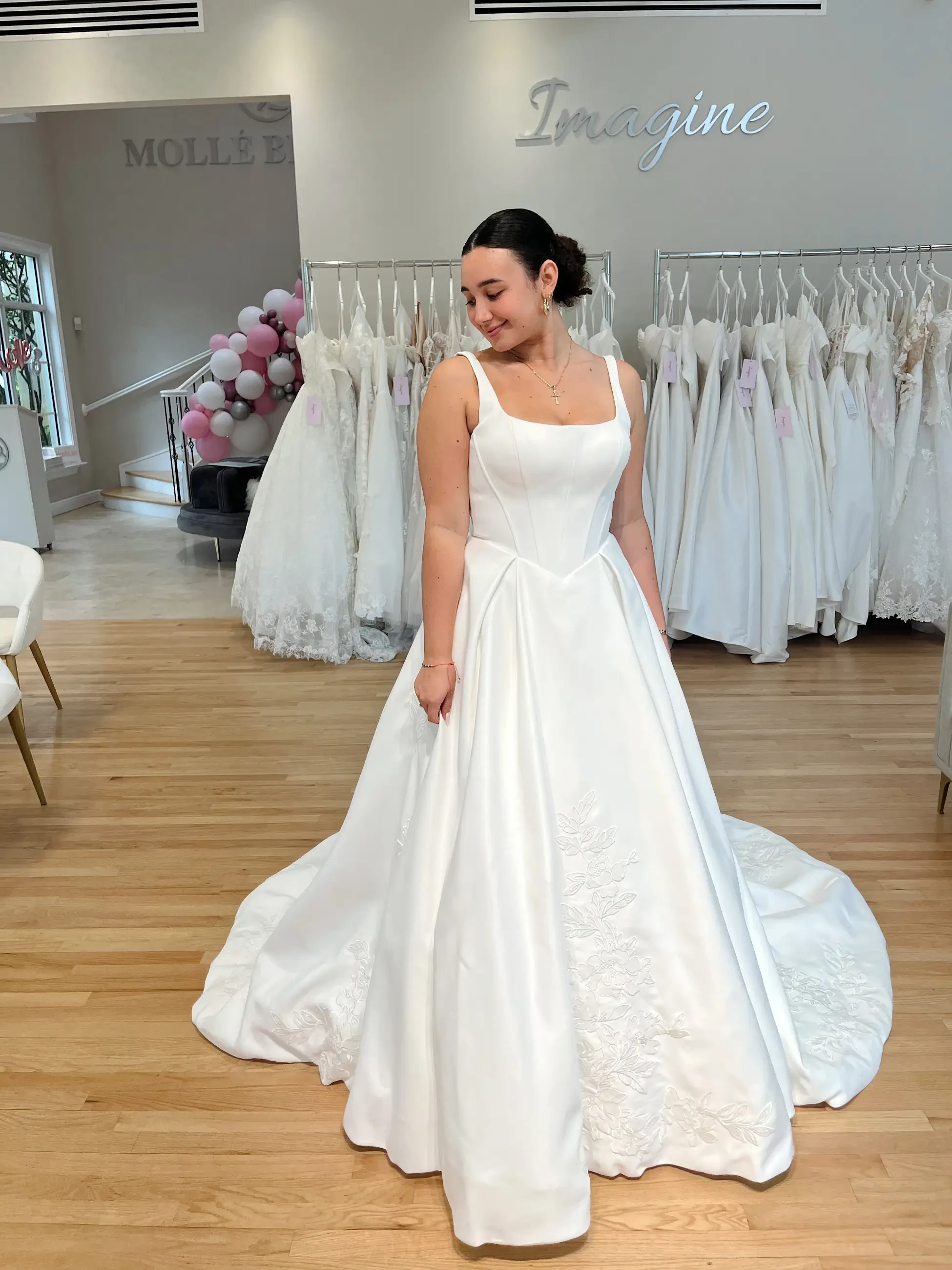 A woman smiles while trying on a white, floral-embroidered wedding dress in a boutique. Gowns hang in the background, creating a joyful, bridal ambiance.