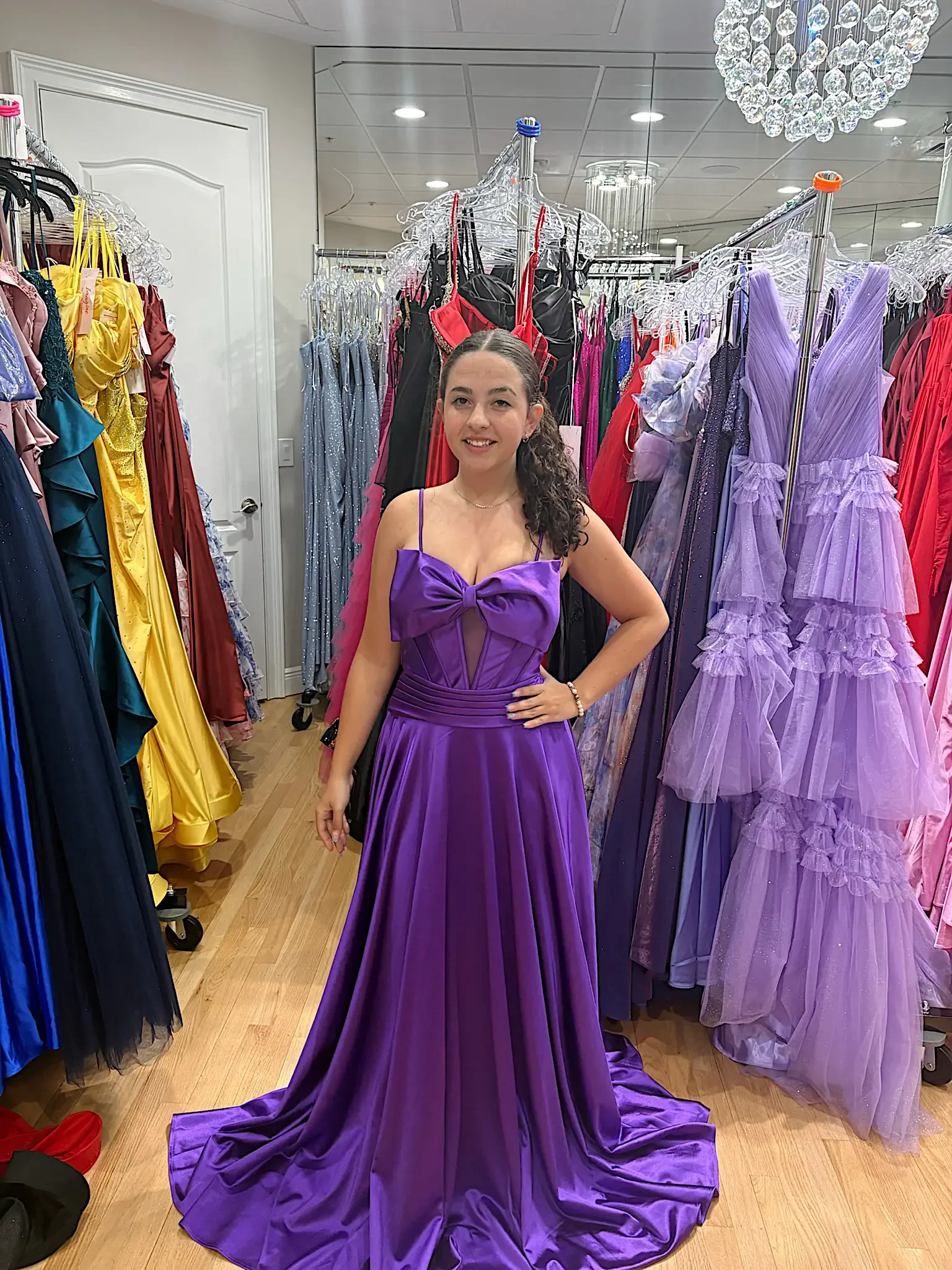 A woman is standing in a dress store, wearing a purple gown with a bow detail on the front. Various colorful dresses are hanging in the background.