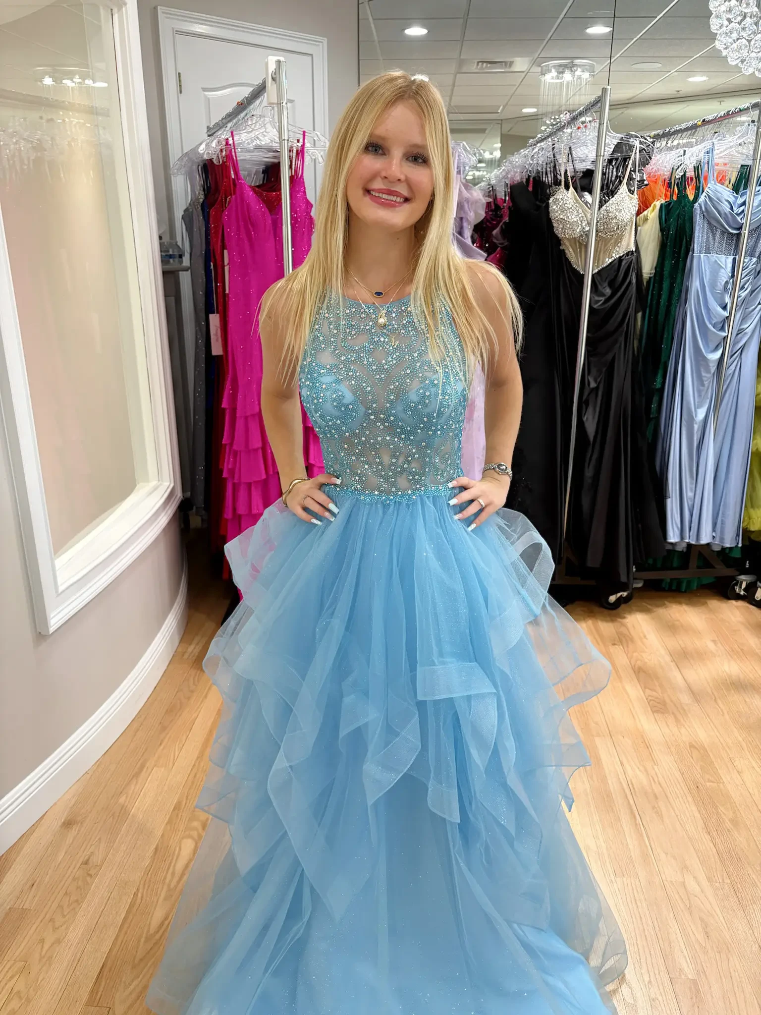 A young woman is smiling, wearing a light blue, beaded gown with a layered tulle skirt, standing in a boutique. Behind her are various colorful dresses.
