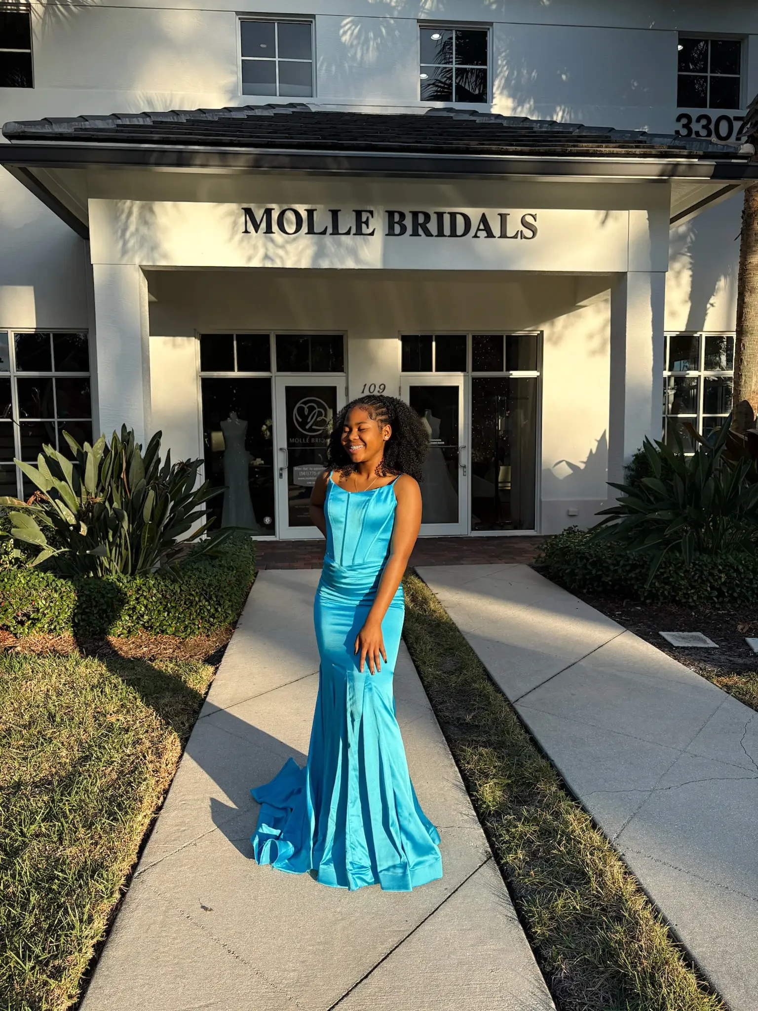 A woman in a blue evening gown stands smiling in front of a bridal shop entrance. She is bathed in sunlight, creating a joyful and elegant atmosphere.