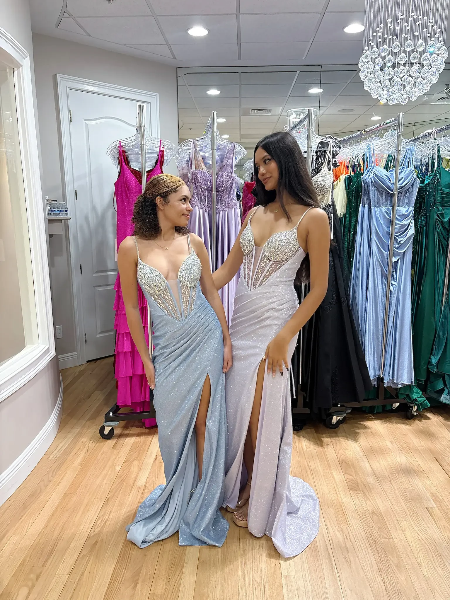 Two women in a boutique wear glamorous, fitted gowns with intricate sparkling details and high slits. They're smiling at each other, surrounded by an array of colorful dresses on racks, creating an elegant and joyful atmosphere.