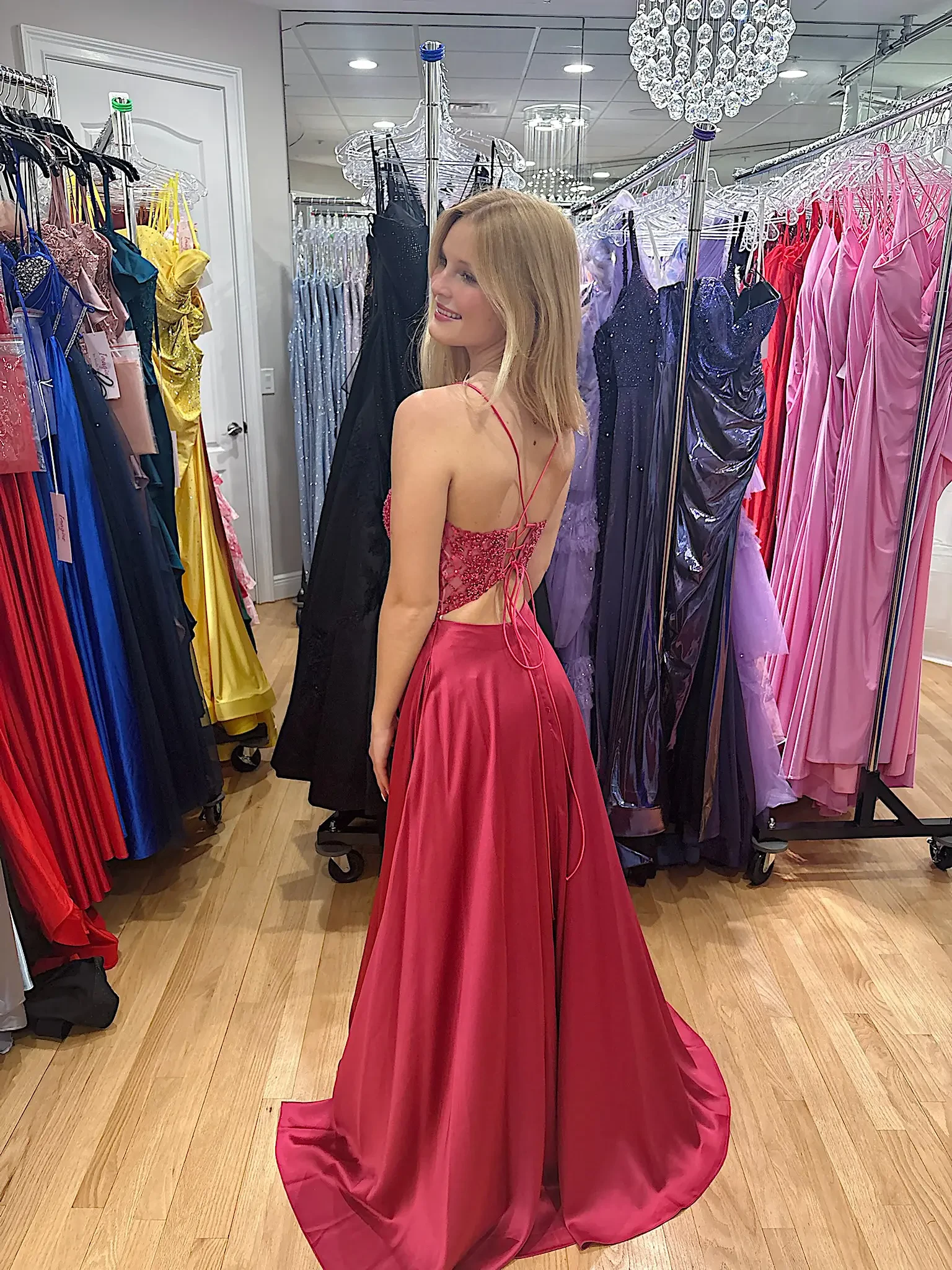 A girl stands in a dress shop, wearing a pink evening gown and looking back over her shoulder. The store features various dresses in the background, hanging on racks.