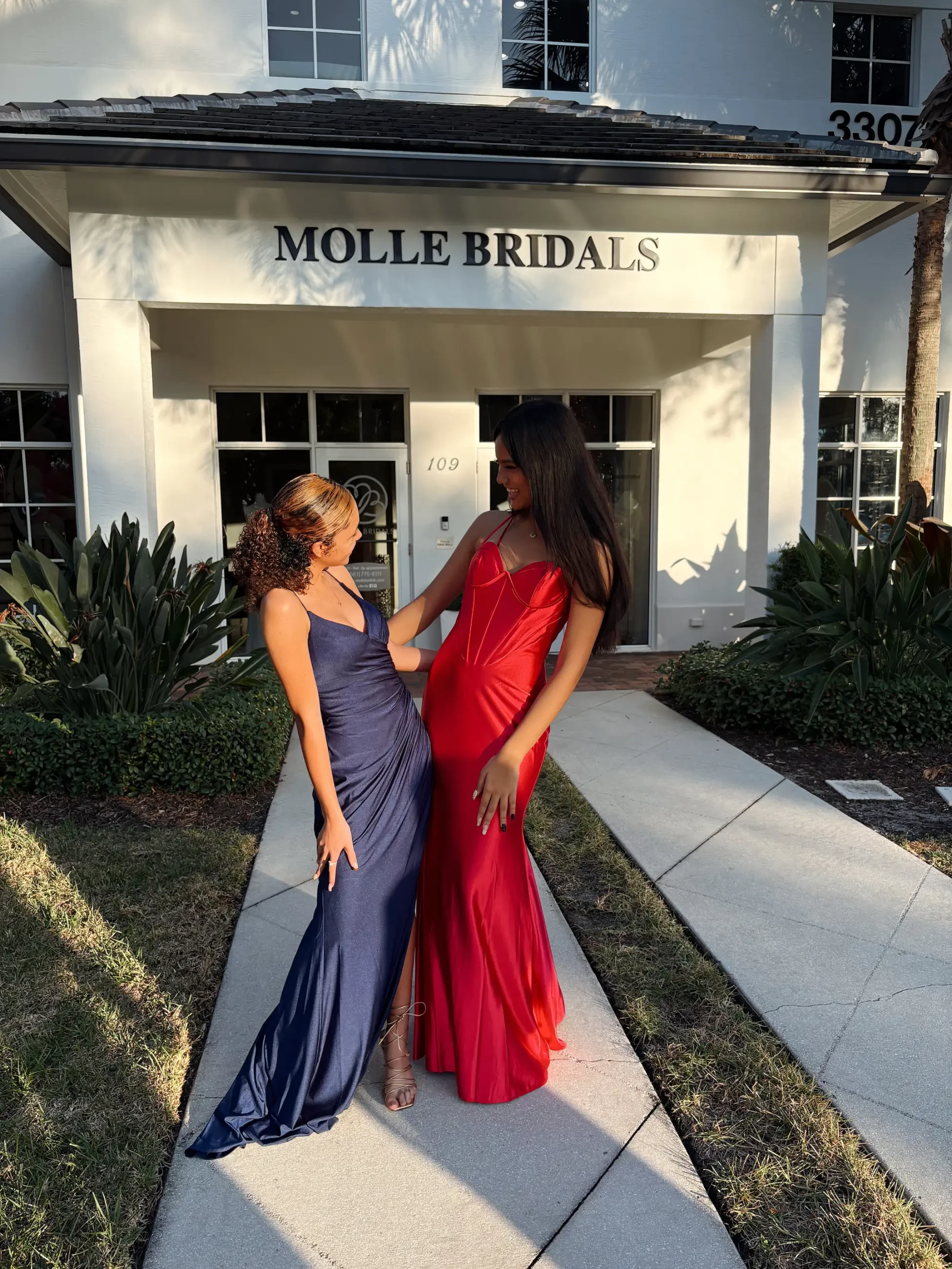 Two women in elegant gowns, one in blue and the other in red, stand happily in front of a bridal store. They smile at each other, radiating joy.