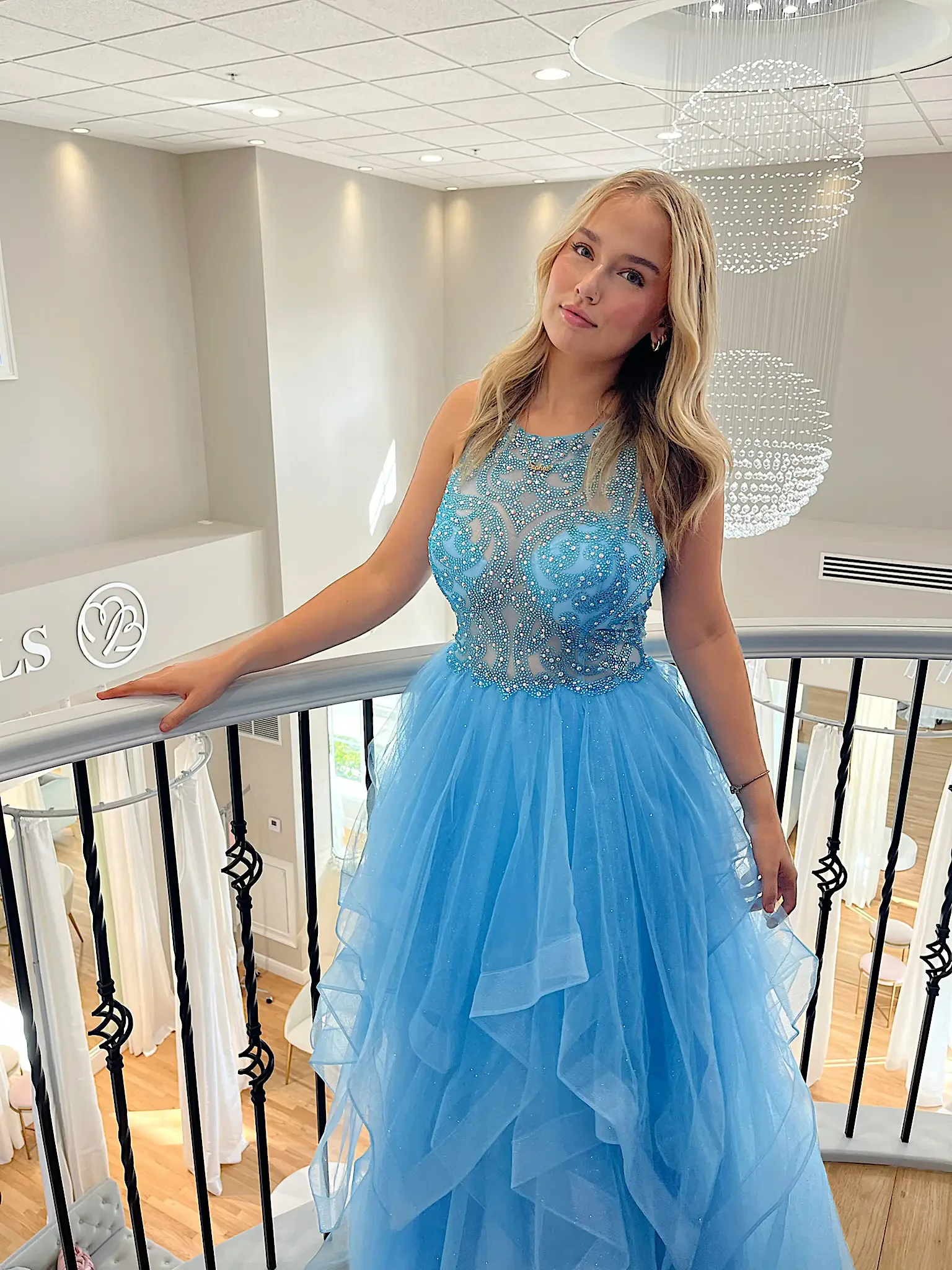 A person in a blue formal dress with layers and embellishments stands near a railing in a bright, elegant space.