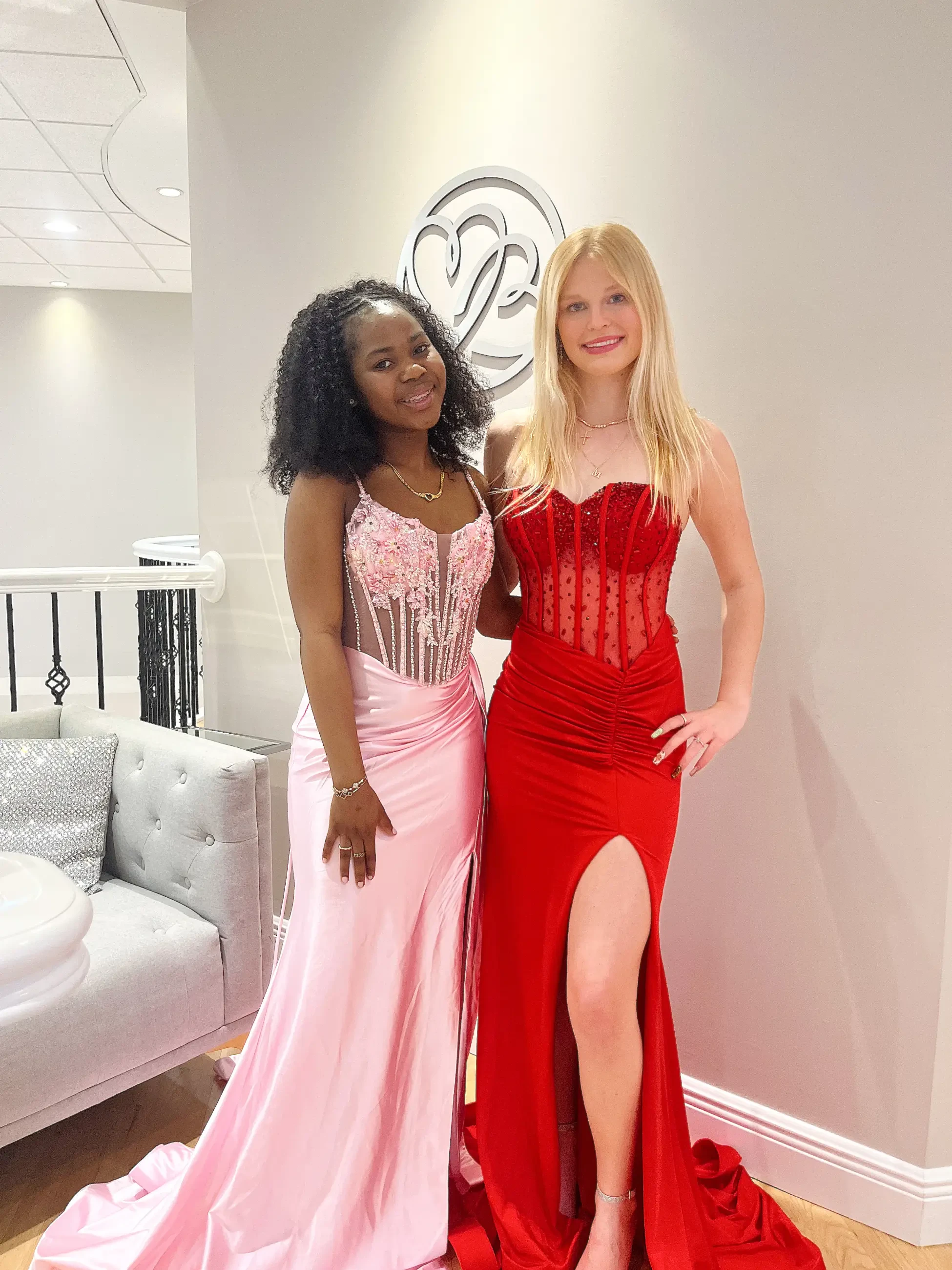 Two women smiling and posing in elegant gowns; one in a pink dress with floral details, the other in a red dress with a thigh-high slit. They stand in a softly lit room with a silver wall decoration, exuding a joyful, celebratory mood.