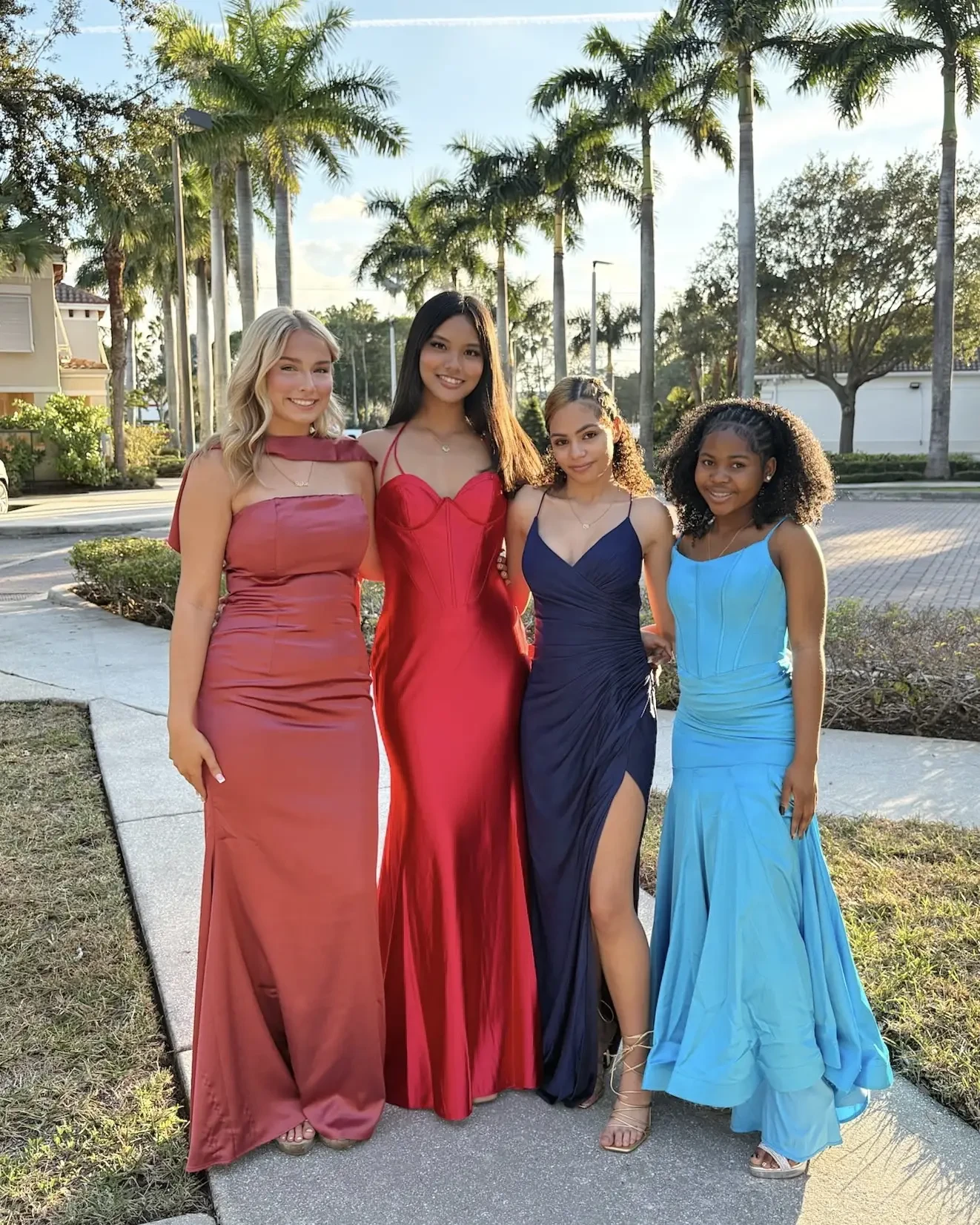 Four young women wearing elegant evening gowns in red, maroon, navy, and turquoise stand outdoors, smiling, with palm trees in the background.