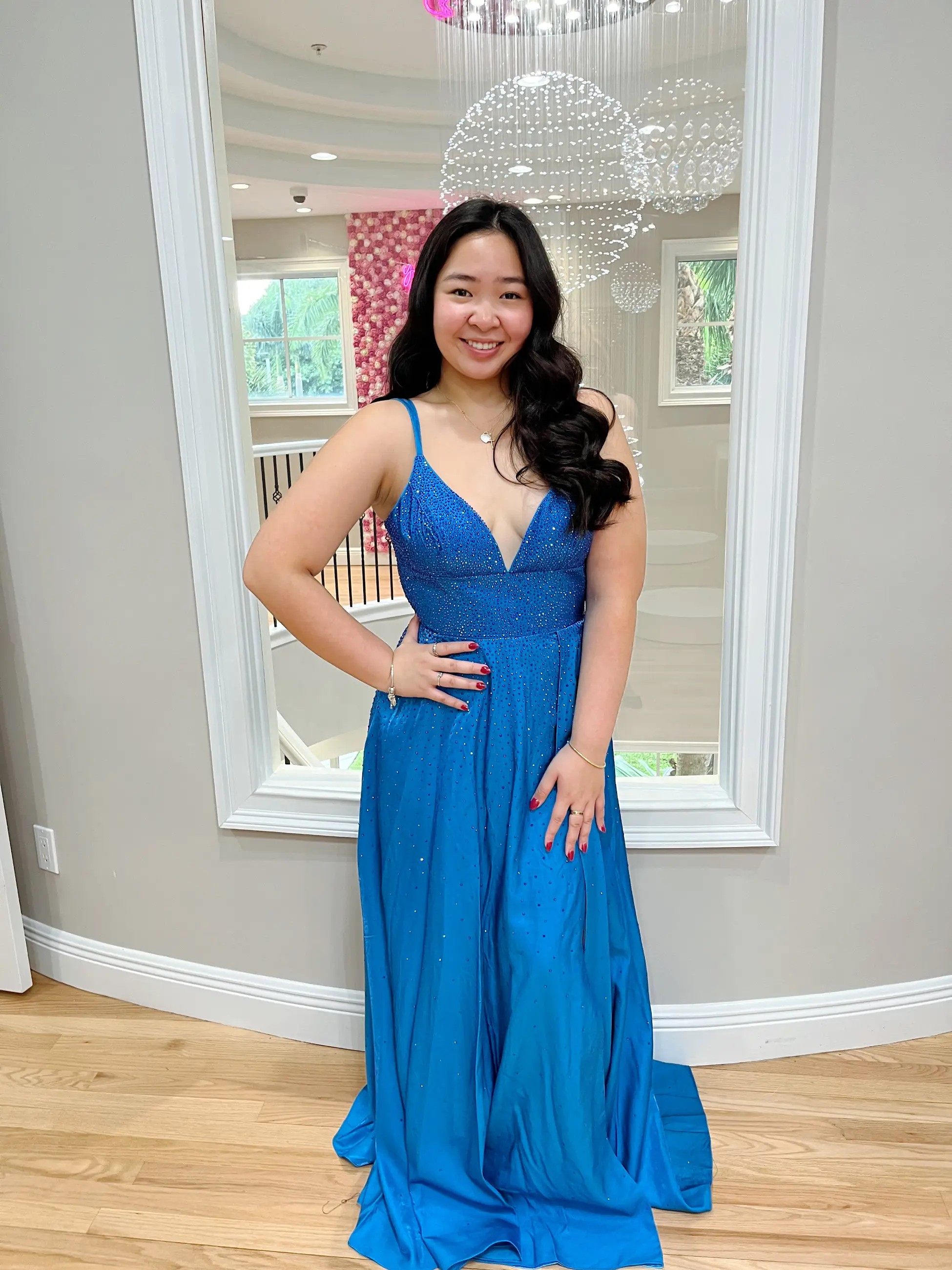 Model is wearing an a-line sleeveless evening gown that’s blue and glittery with a plunging neckline