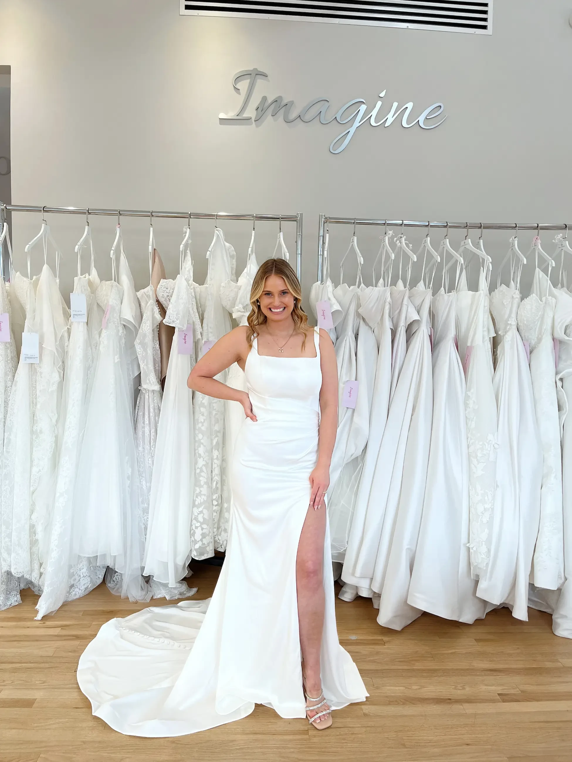 A woman in a white wedding dress with a thigh-high slit poses in front of a rack of white dresses.