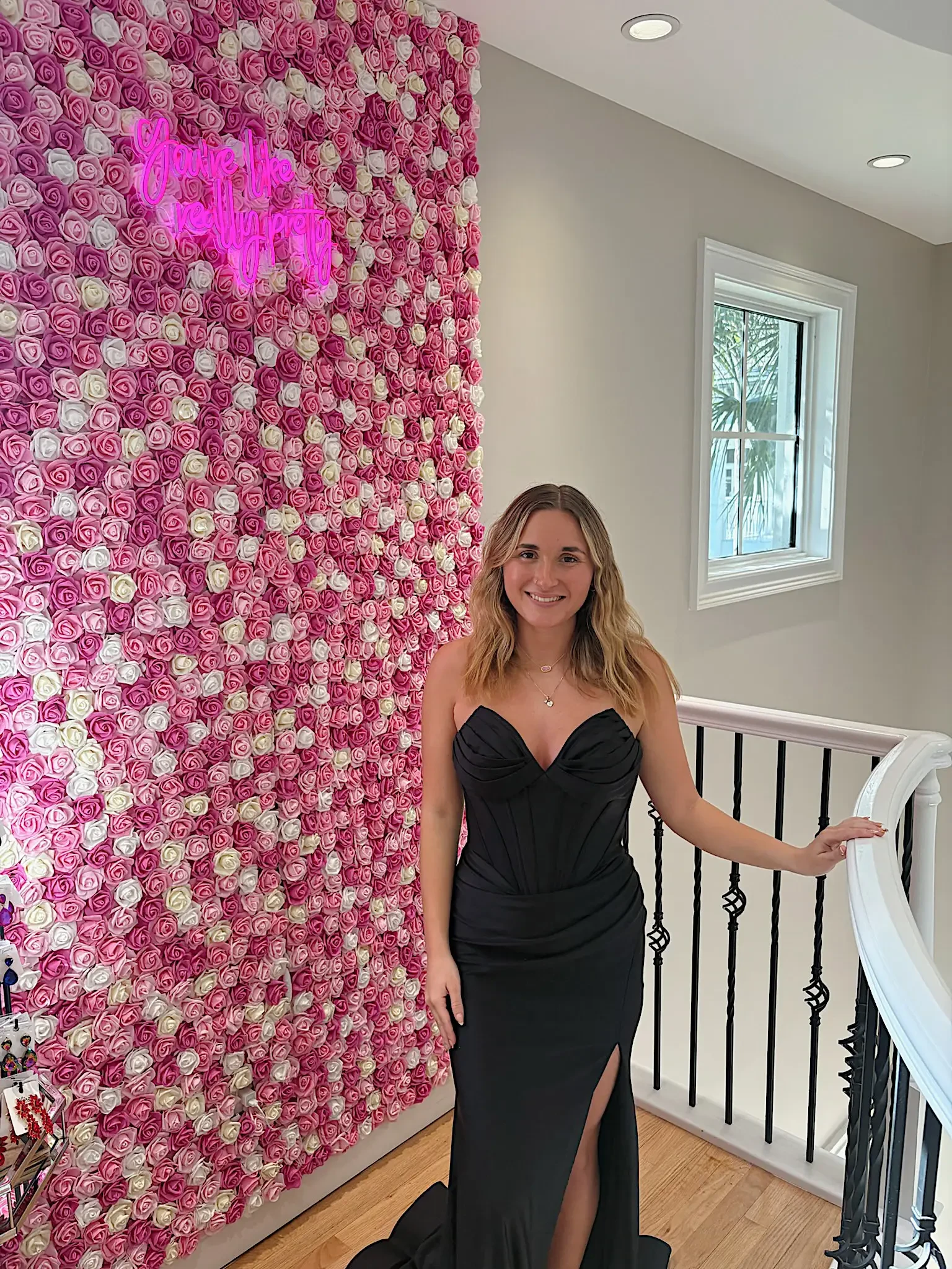A woman standing next to a wall covered in pink and white roses, wearing a black dress with a high slit.