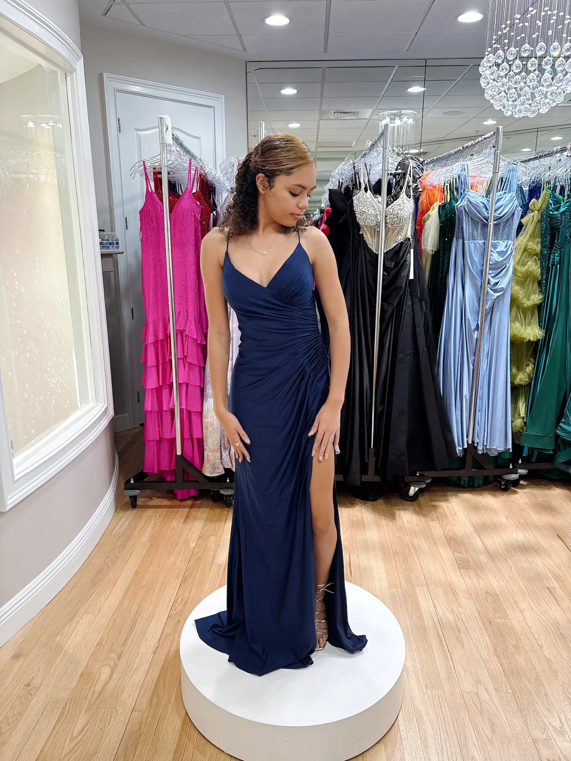 A woman in a navy blue gown with a high slit stands on a round platform in a boutique. She is surrounded by colorful dresses, conveying a sense of elegance.