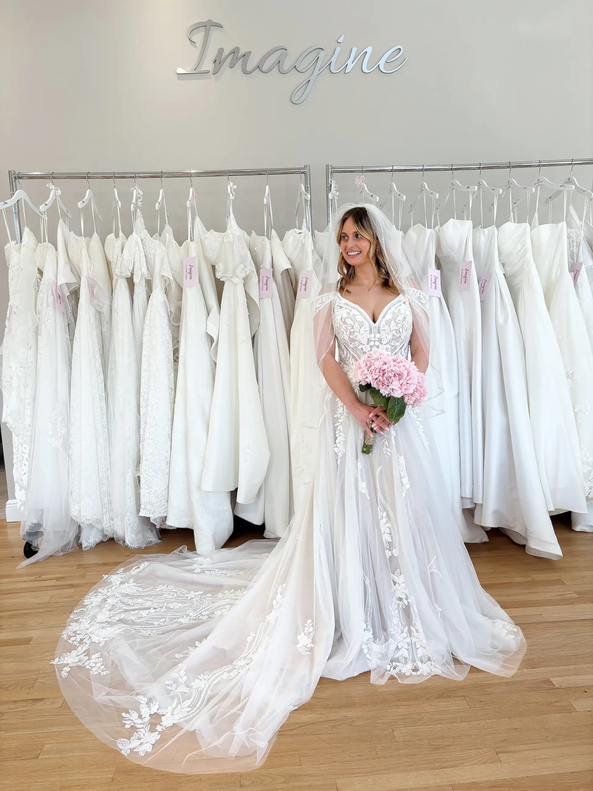 A bride stands in front of a rack of white wedding dresses, wearing a gown with a long train and lace details, holding pink flowers, exuding joy under the word 