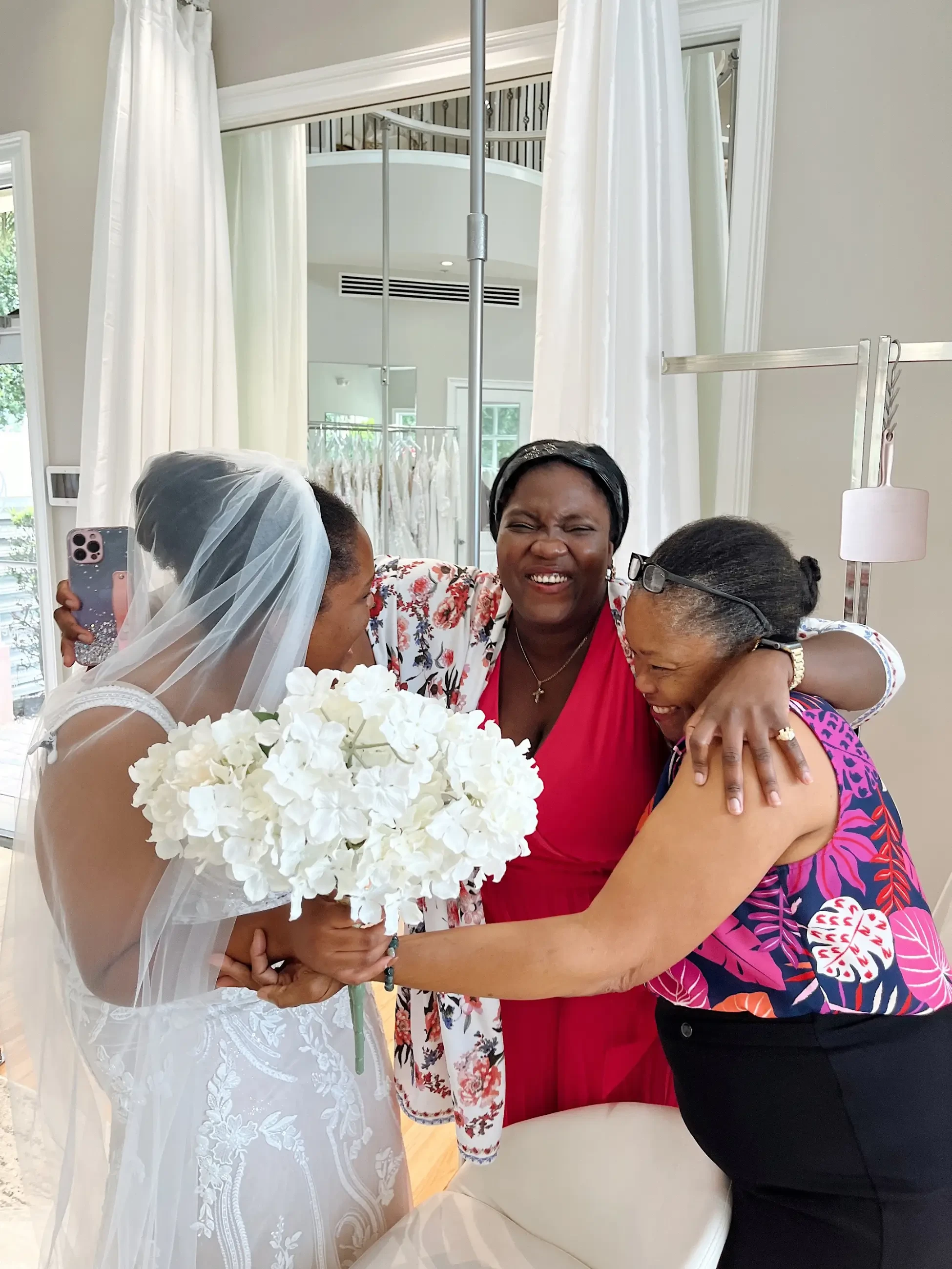 A bride in a lace wedding dress and veil embraces two smiling women. One holds white flowers. The scene is joyful and intimate, framed by light curtains.