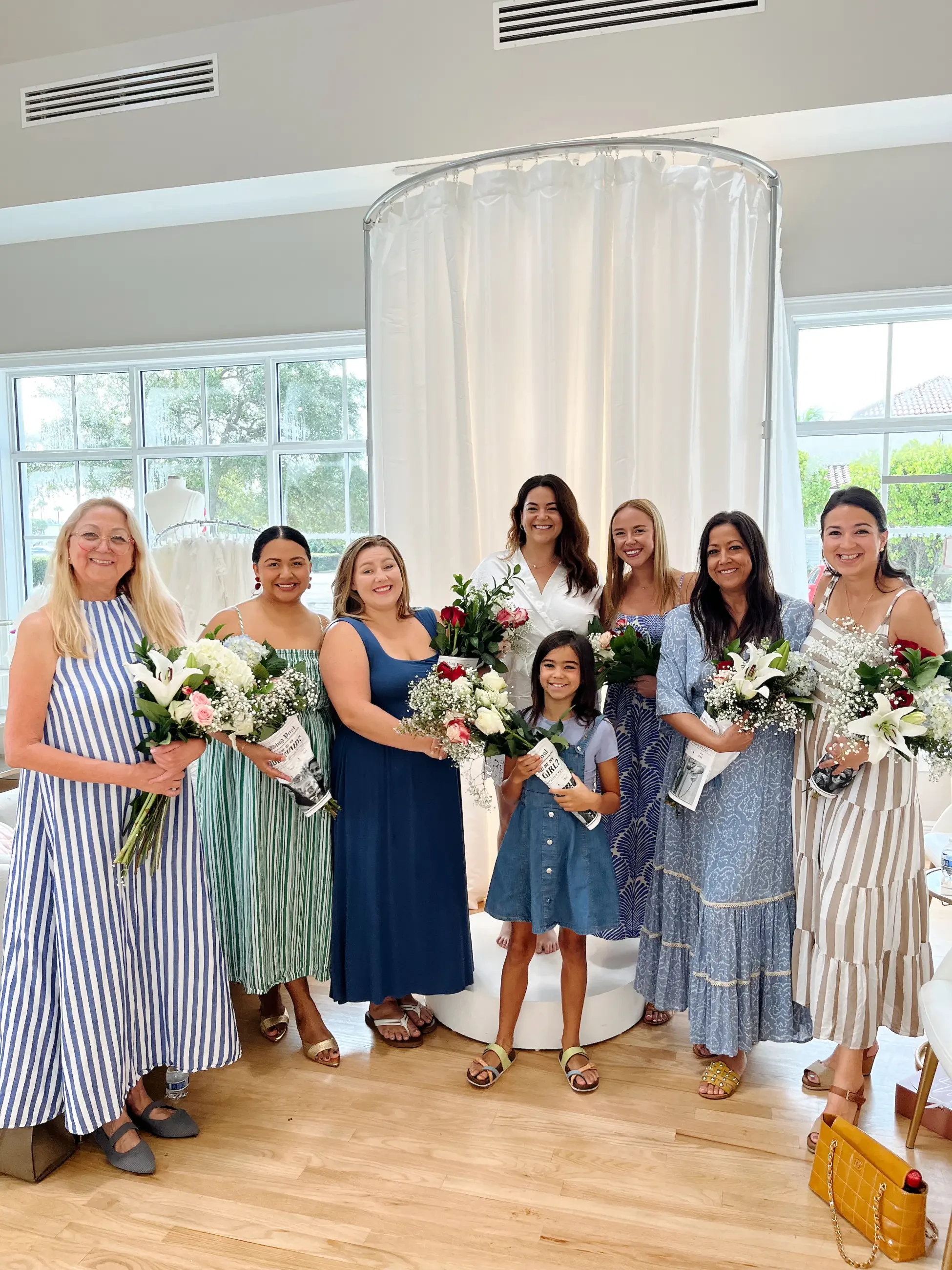 A group of seven women and one girl stand smiling, holding bouquets with white and pink flowers in a bright room with wooden floors and large windows.