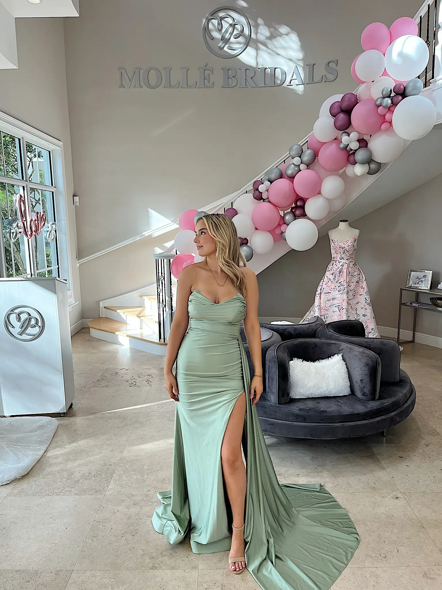 A woman wearing a light green gown with a slit stands in a bridal shop, featuring decorative balloons and wedding attire in the background.