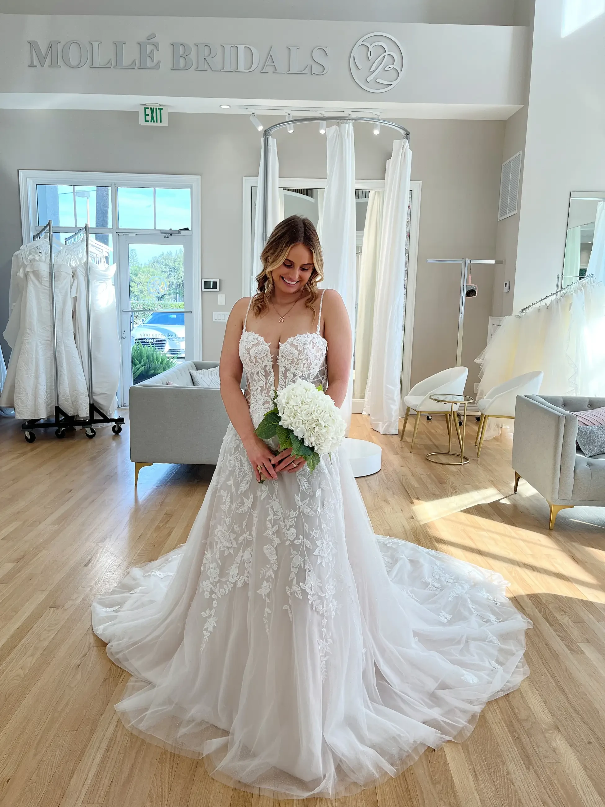 Bride in a lace gown holds white flowers, smiling in a bright bridal store. The elegant dress features intricate embroidery and a long train.