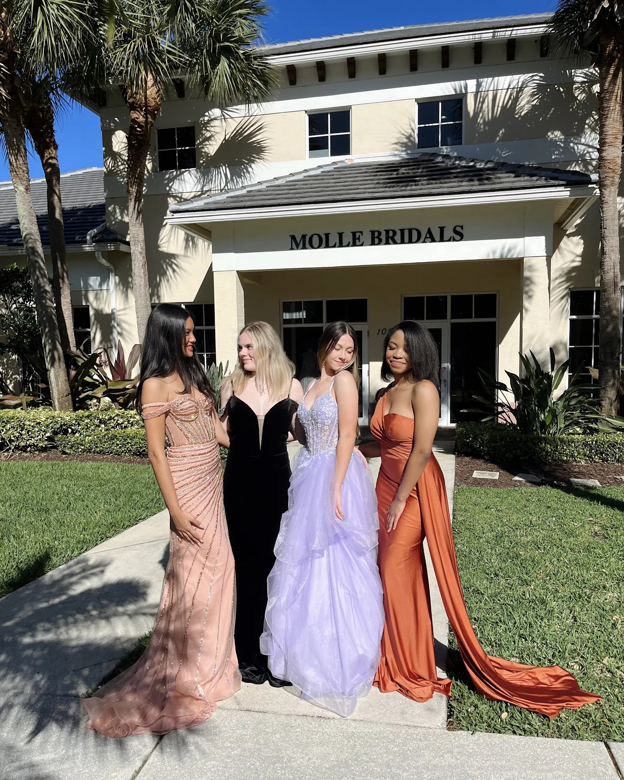 In this photo, there are four models smiling and laughing with each other wearing prom/evening gowns (one in champagne, one in black, one in purple, and one in orange)
