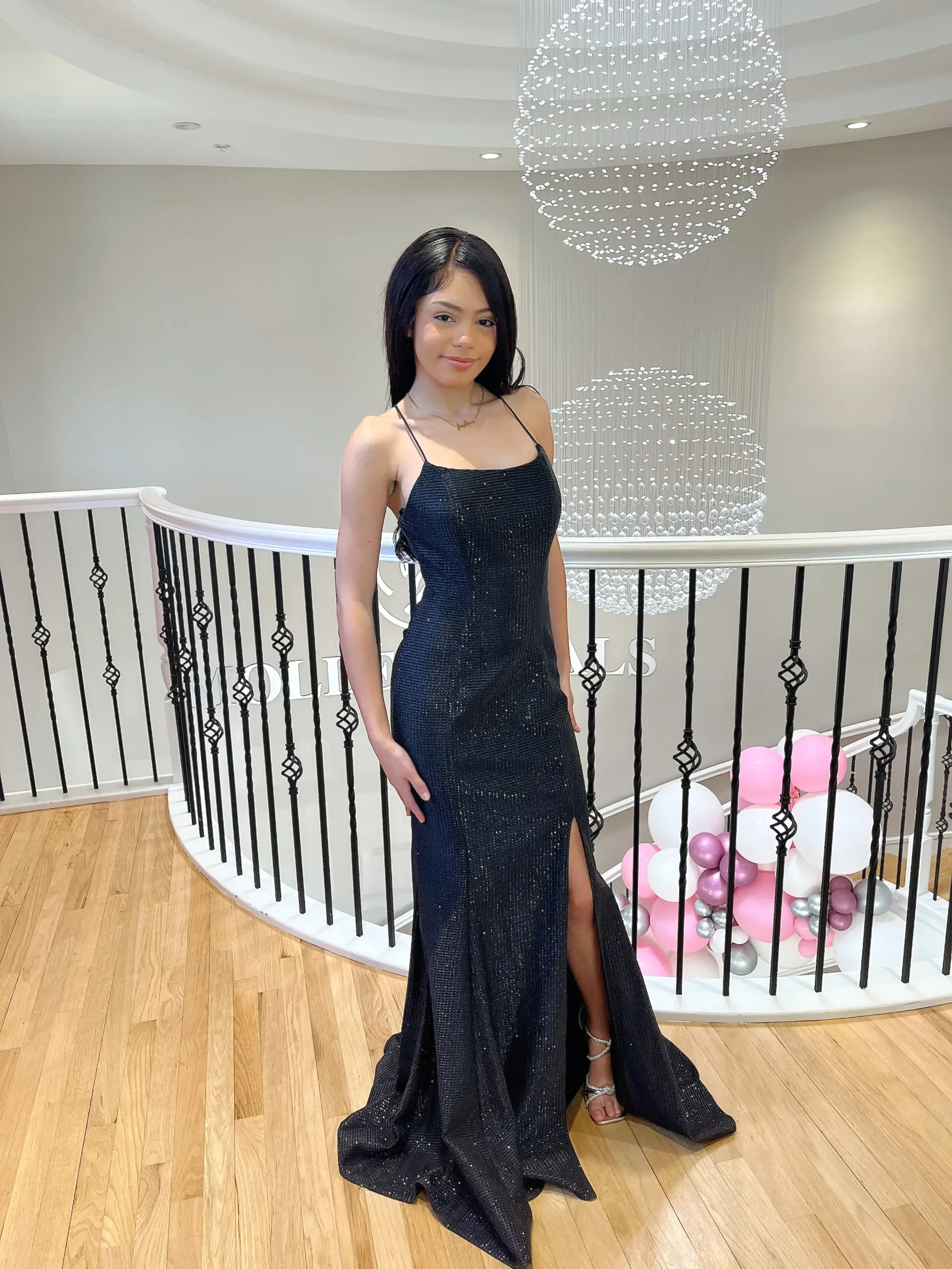 Young woman in a sparkling black gown stands gracefully by a curved railing. Behind her, a chic chandelier and pink and white balloons add an elegant touch.