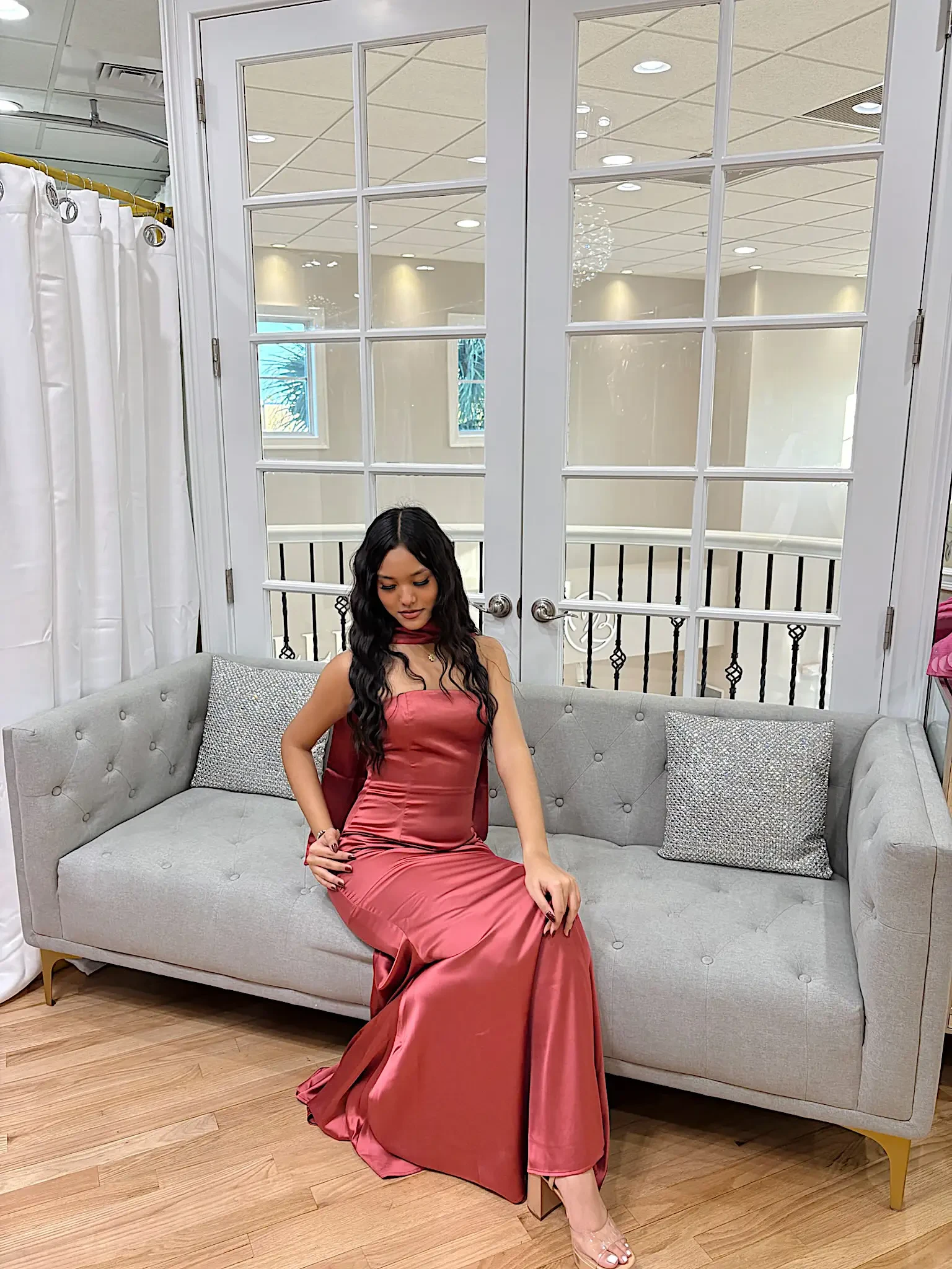 A woman in a pink satin dress sitting on a gray sofa in a well-lit room.
