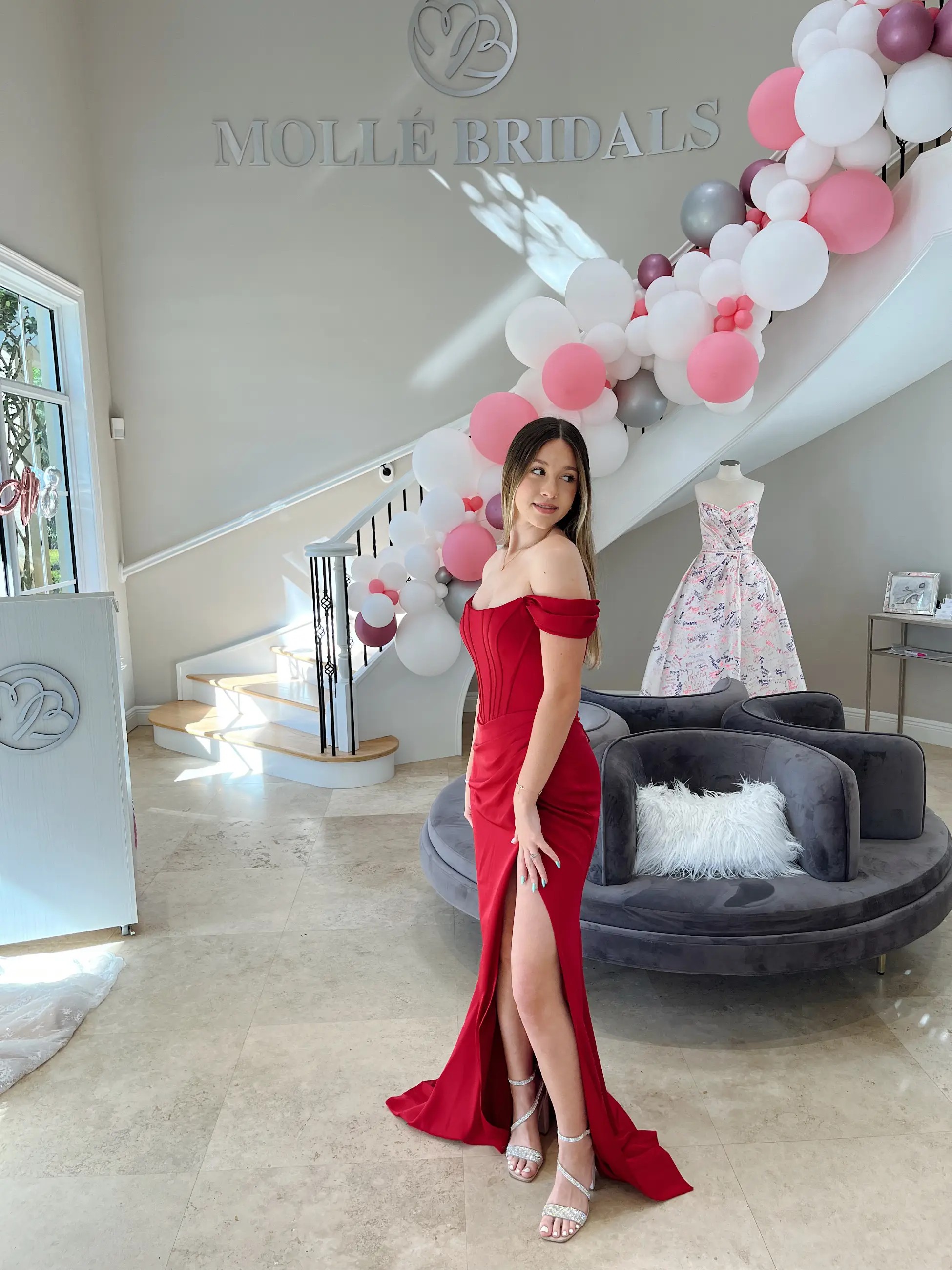 Model is wearing a fitted red evening gown with an off the shoulder neckline and high slit