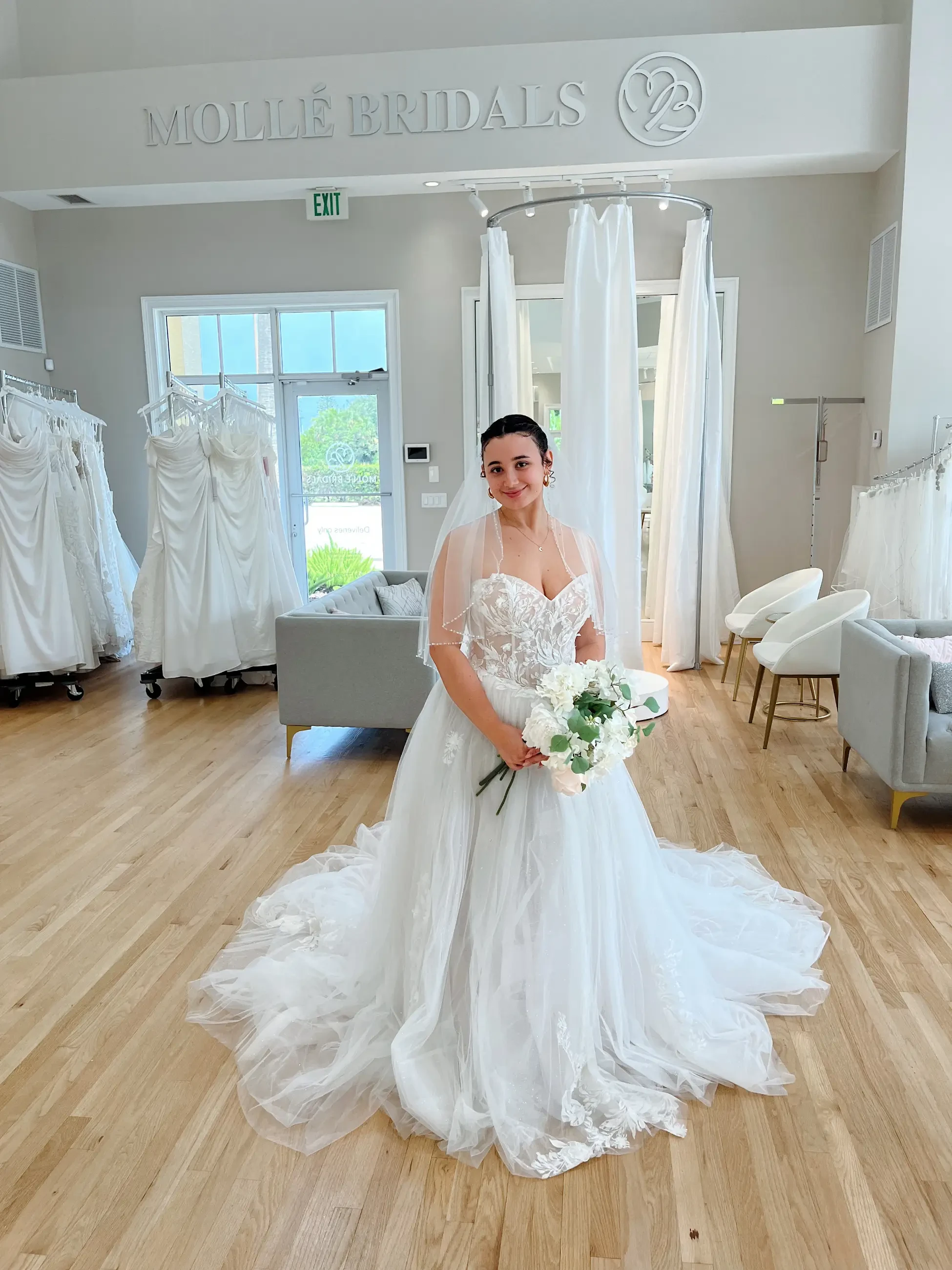 A bride in a lace wedding gown holds a bouquet, smiling in a bridal shop. The room is elegant with wooden floors and displayed dresses, creating a joyful atmosphere.