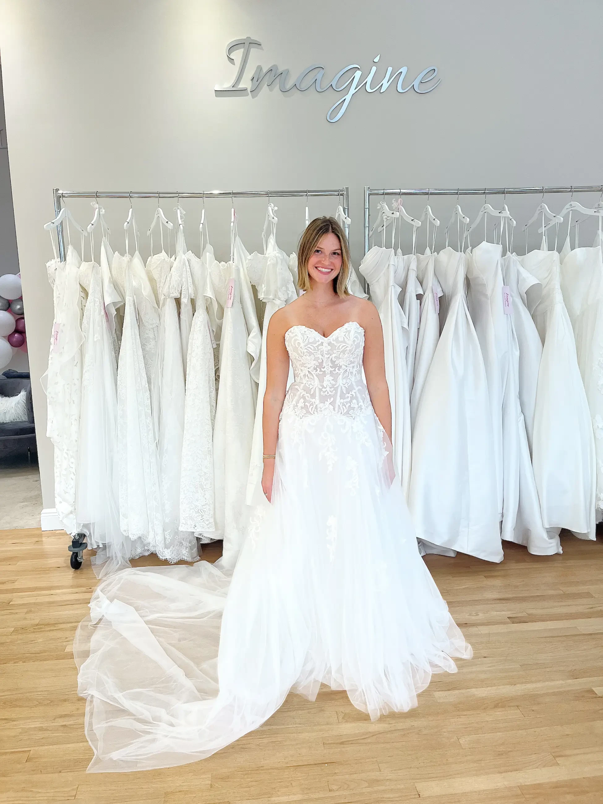 A woman is smiling and standing in a bridal boutique, wearing a beautiful wedding dress with intricate lace detailing. In the background, there are several white wedding dresses hanging on racks.