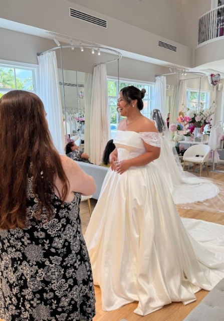 A bride in a white wedding dress is standing in front of a mirror, while a woman in a black and white dress looks on. The setting appears to be a bridal shop.
