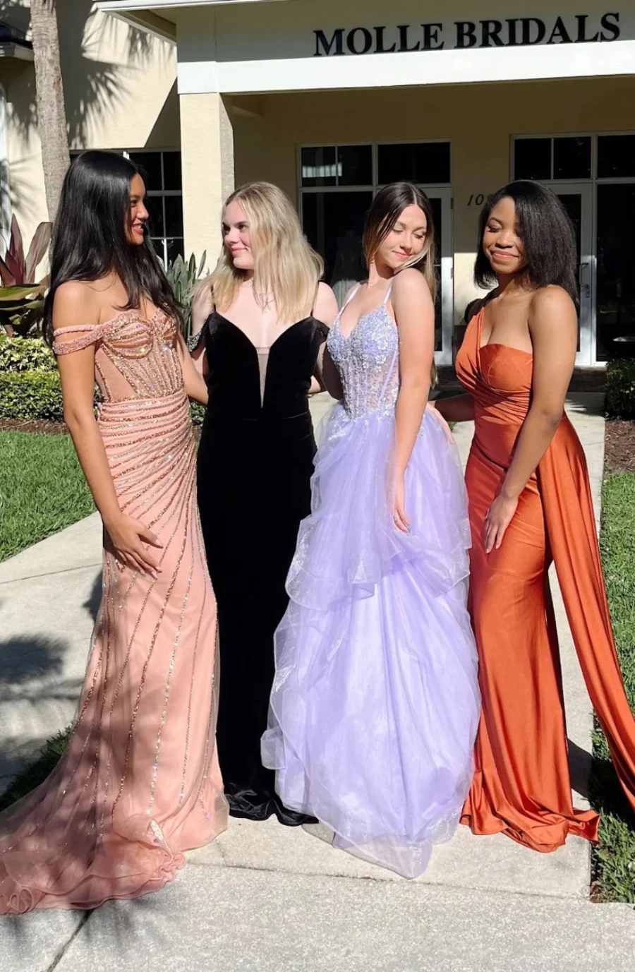four models wearing floor length gowns, in these colors: champagne, black, light purple, and orange