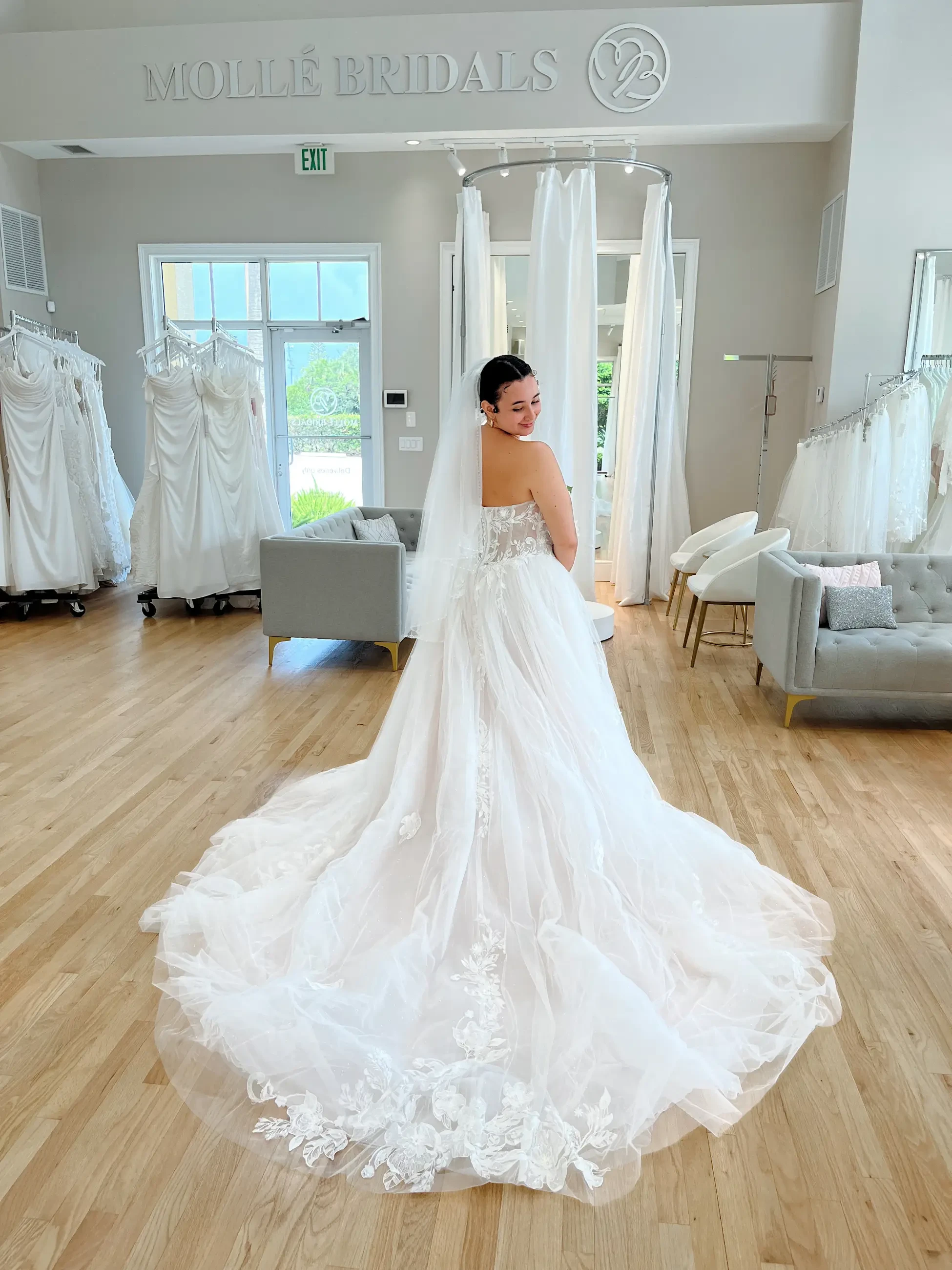 Bride in a flowing white gown with lace details stands in a bridal boutique. The room has wooden floors, elegant sofas, and racks of dresses.