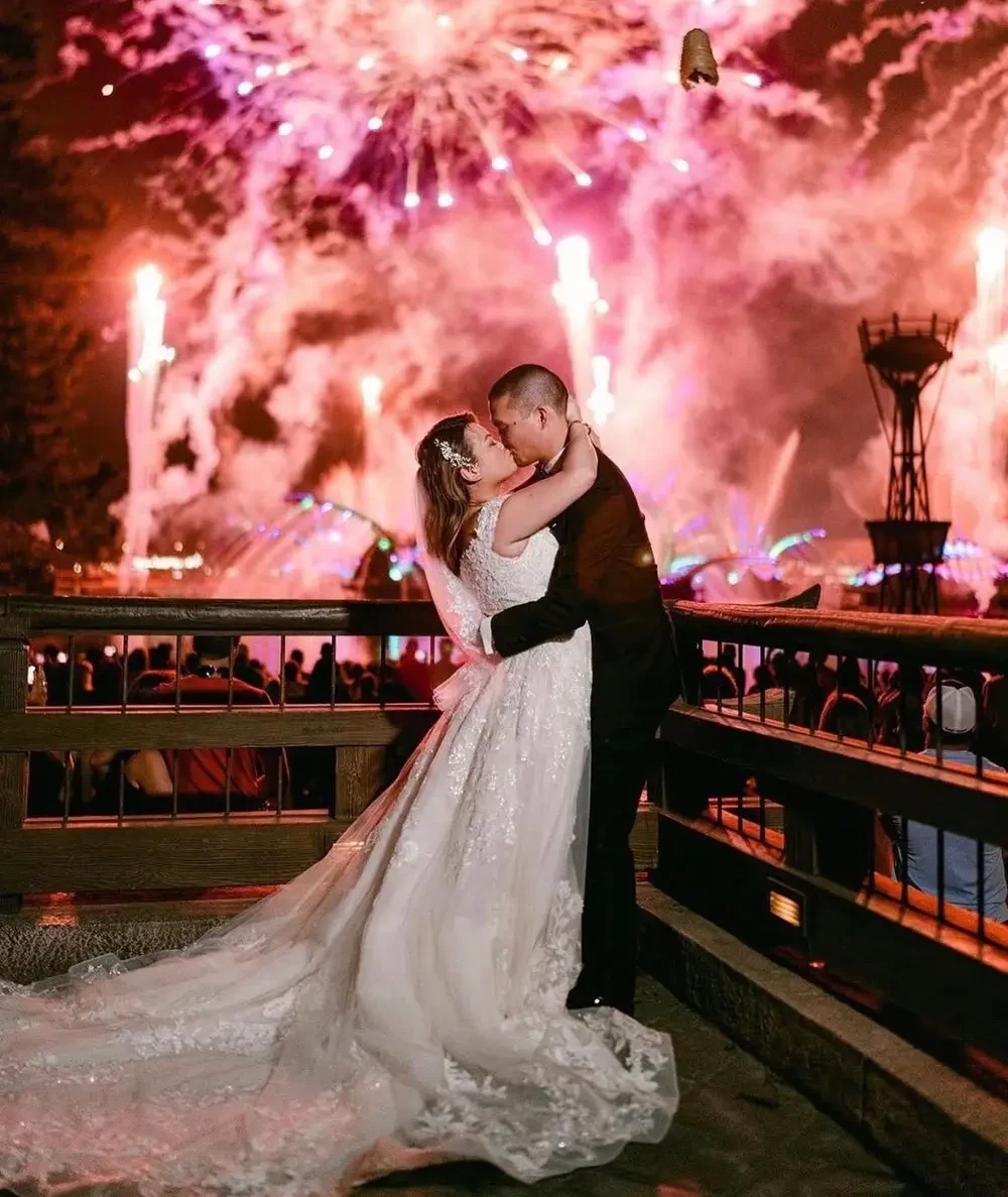 A couple in formal attire embraces in front of a spectacular fireworks display.