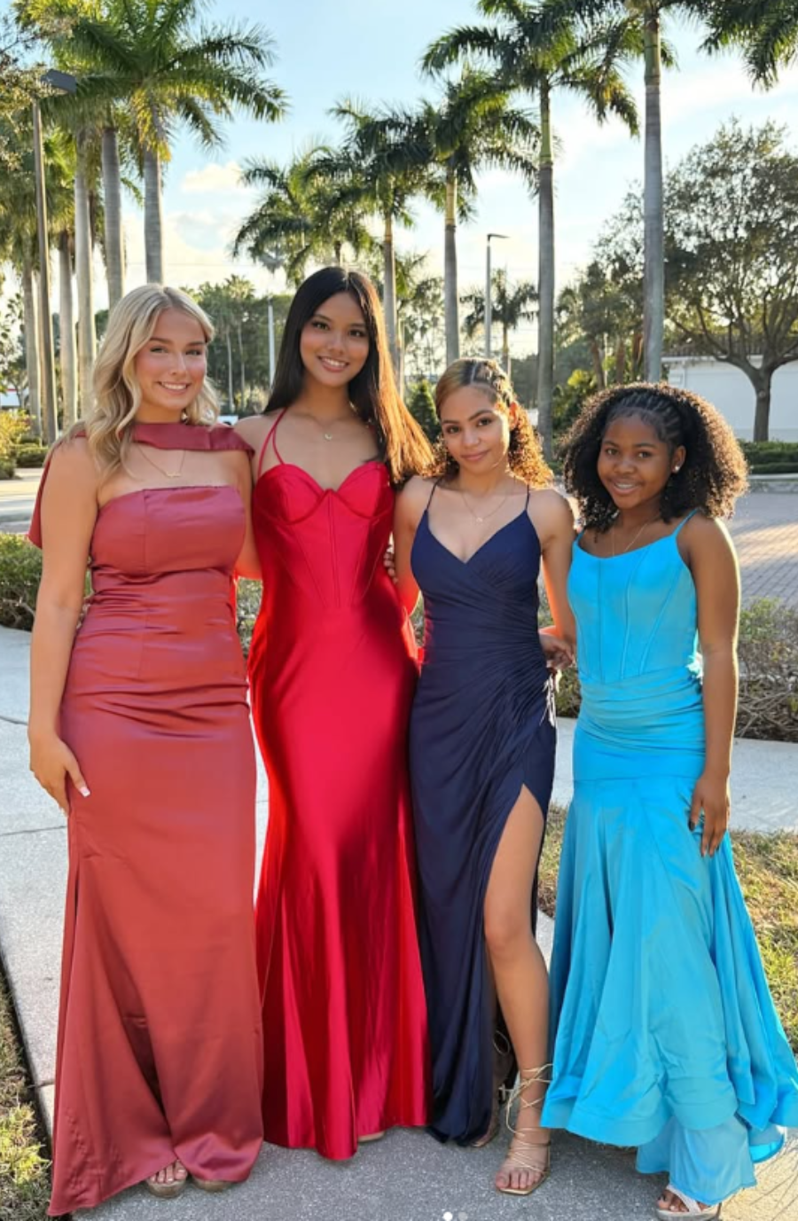 Four young women in colorful evening gowns stand together outdoors, smiling. Palm trees and a sidewalk create a sunny, elegant setting.