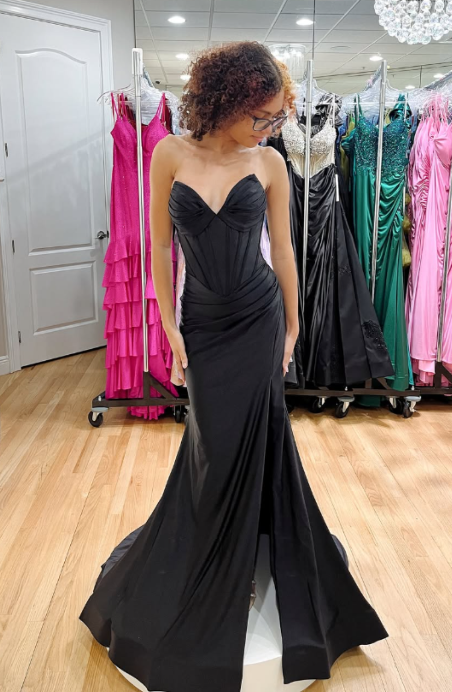 A woman in a fitting room wears a strapless, black mermaid gown, looking down. Bright dresses hang in the background, enhancing the elegant atmosphere.