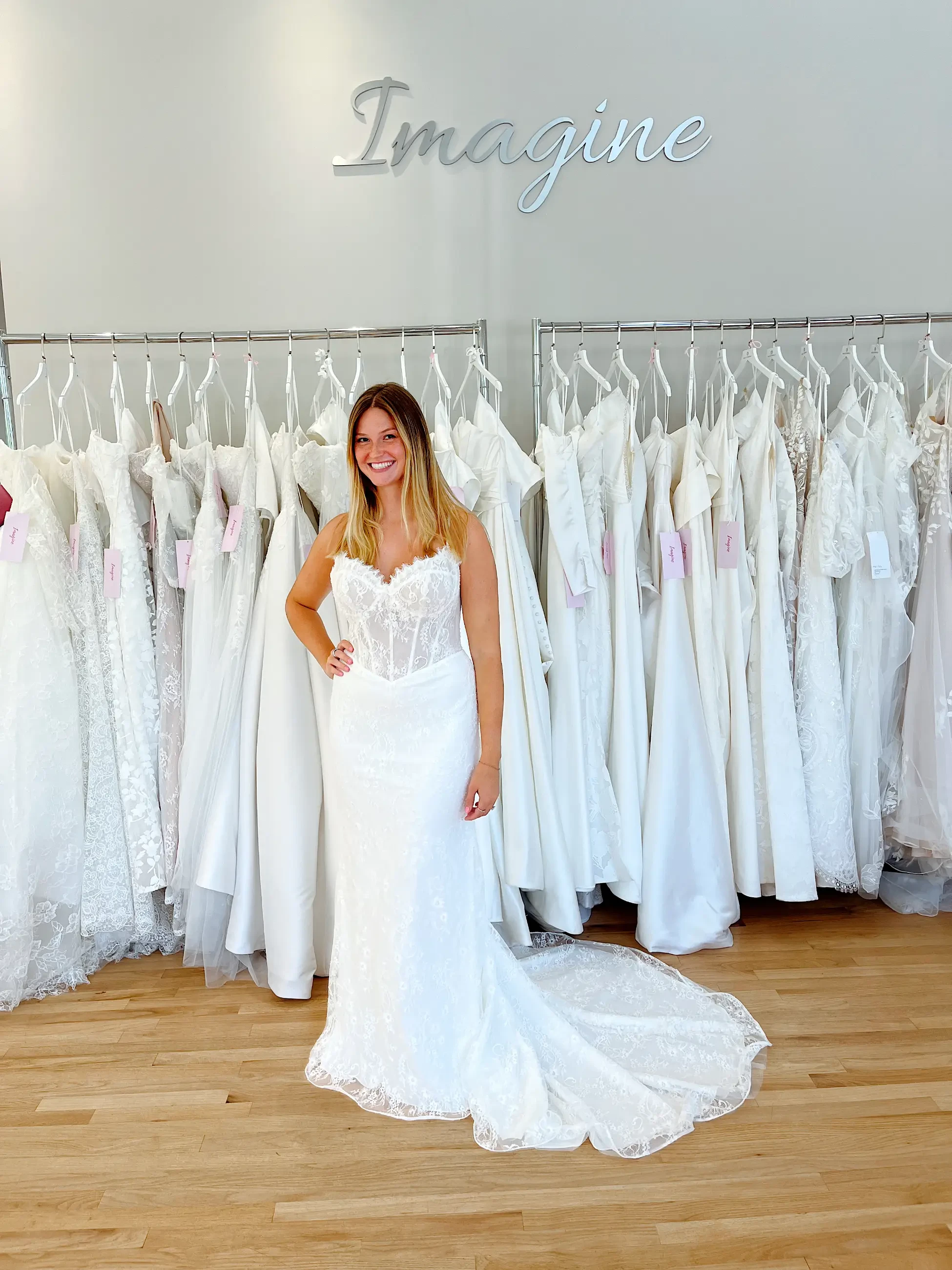 A woman in a white lace wedding dress stands smiling in a bridal shop. Behind her, a display of various wedding gowns hangs on racks. The word 