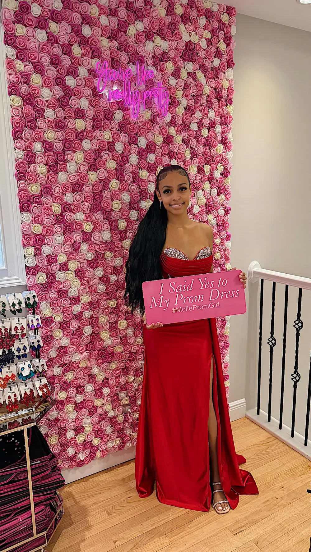 A person in a red dress holding a sign in front of a wall covered in pink and white flowers.