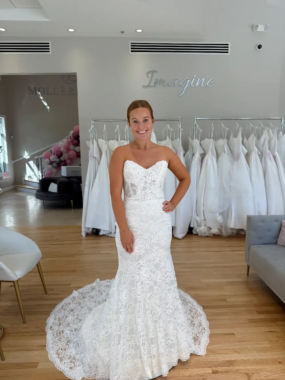 A woman in a white wedding dress poses in a bridal shop surrounded by wedding gowns.
