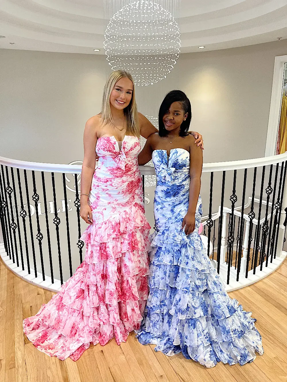 Two women standing on a staircase, wearing elegant, strapless, frilly gowns&mdash;one in pink and the other in blue.