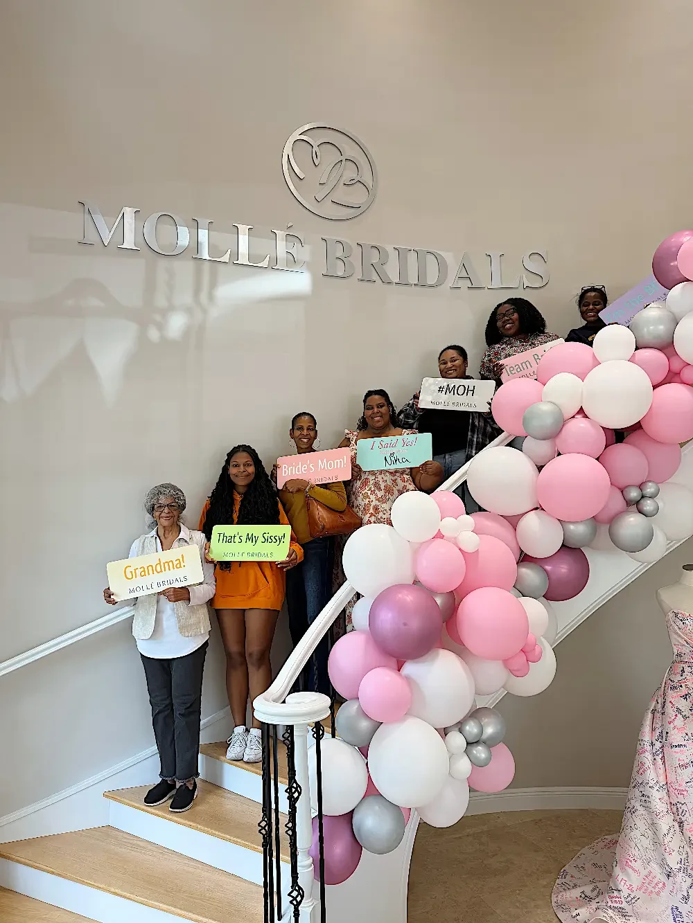 A group of people holding signs at a bridal shop, with balloons decorating the staircase.