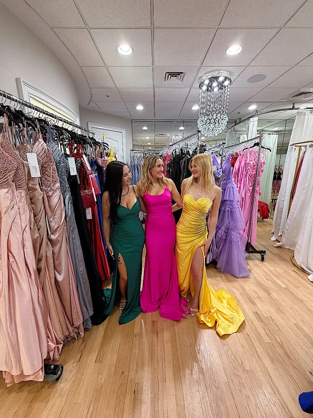 Three women standing together in a clothing store filled with various dresses, each wearing a different colored gown: one in green, one in pink, and one in yellow.