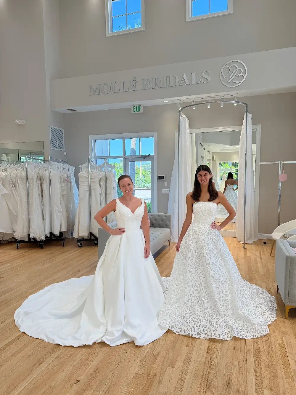 Two brides are trying on wedding dresses in a bridal shop. One is wearing a simple white gown with a train, and the other is in a white dress with intricate lace detailing. The shop has a bright, airy atmosphere with several wedding dresses displayed in the background.